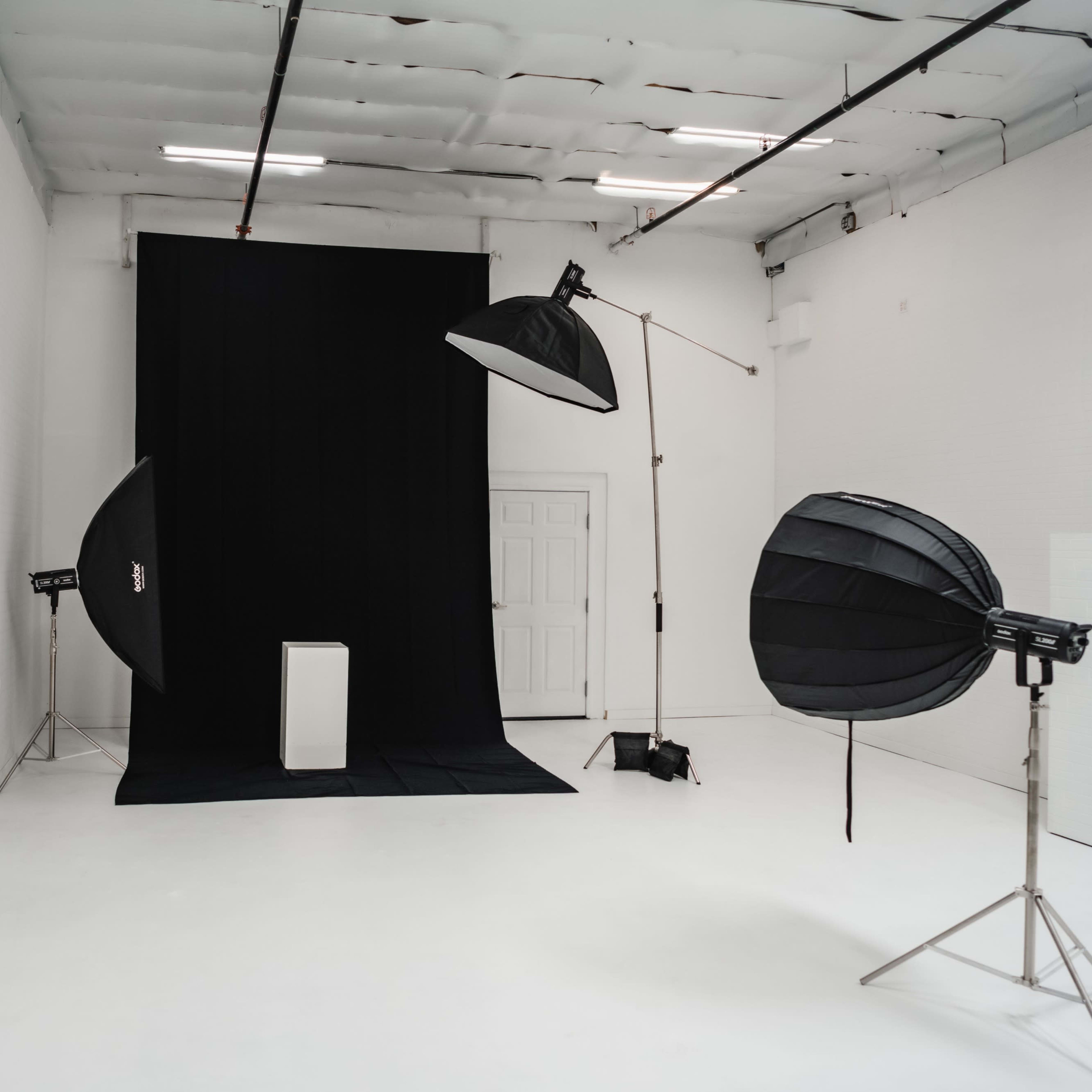 A photo studio features a black backdrop with a white pedestal and two large softbox lights positioned on either side.