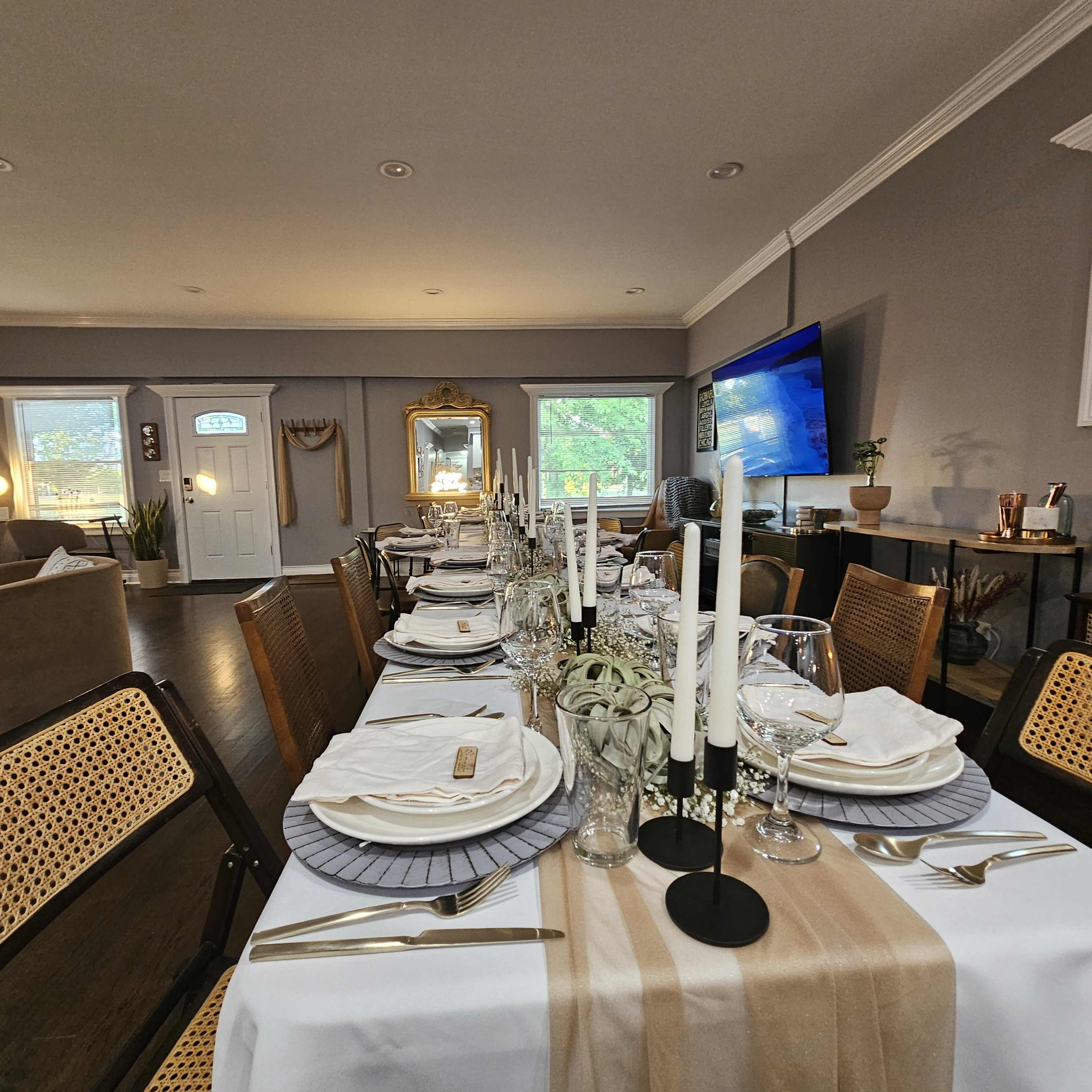A long dining table is elegantly set with white linens, silverware, glassware, and tapered candles, surrounded by a stylishly decorated room featuring a television and large windows.