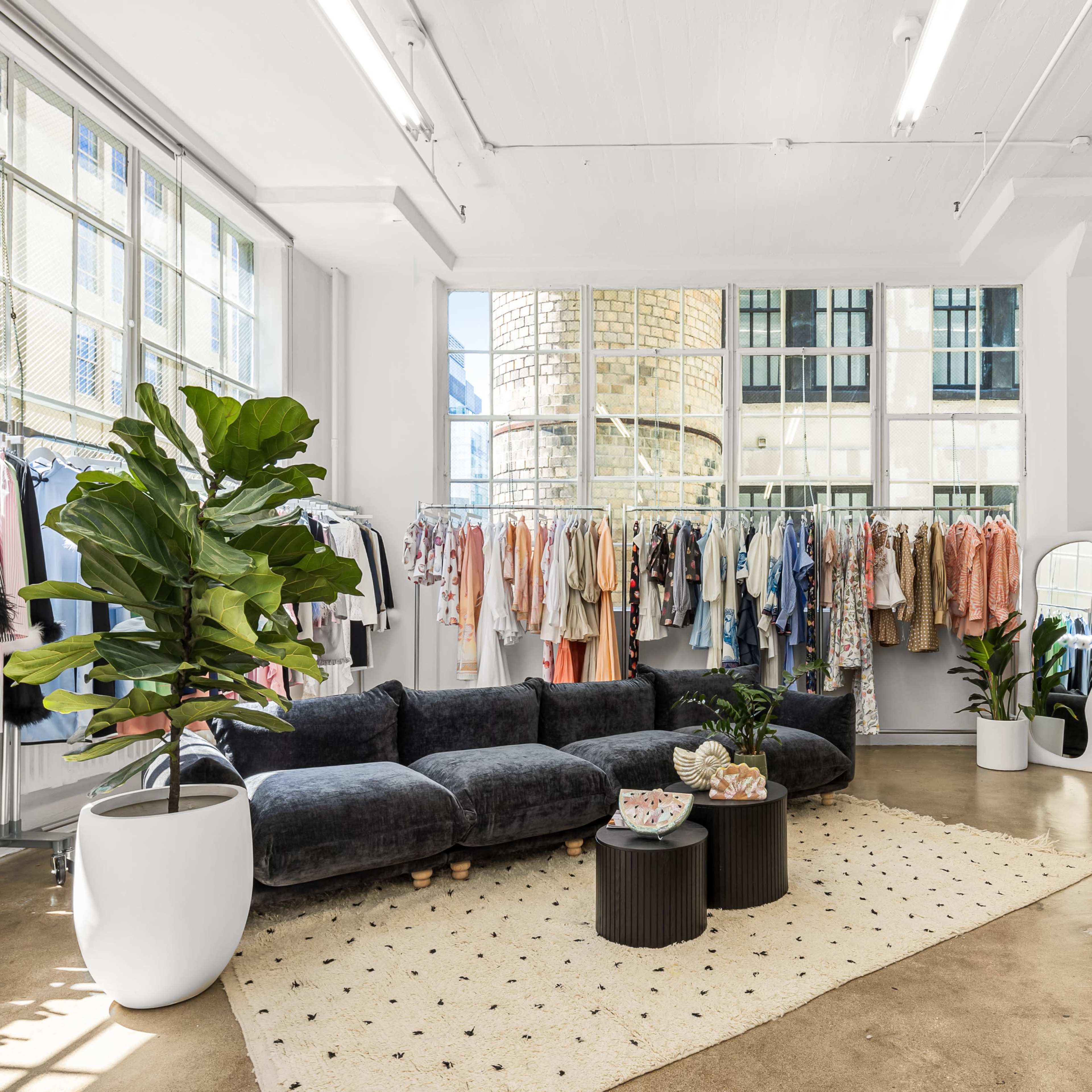 A clothing boutique features racks of apparel against large windows, with a plush seating area and decorative plants arranged on a textured rug.