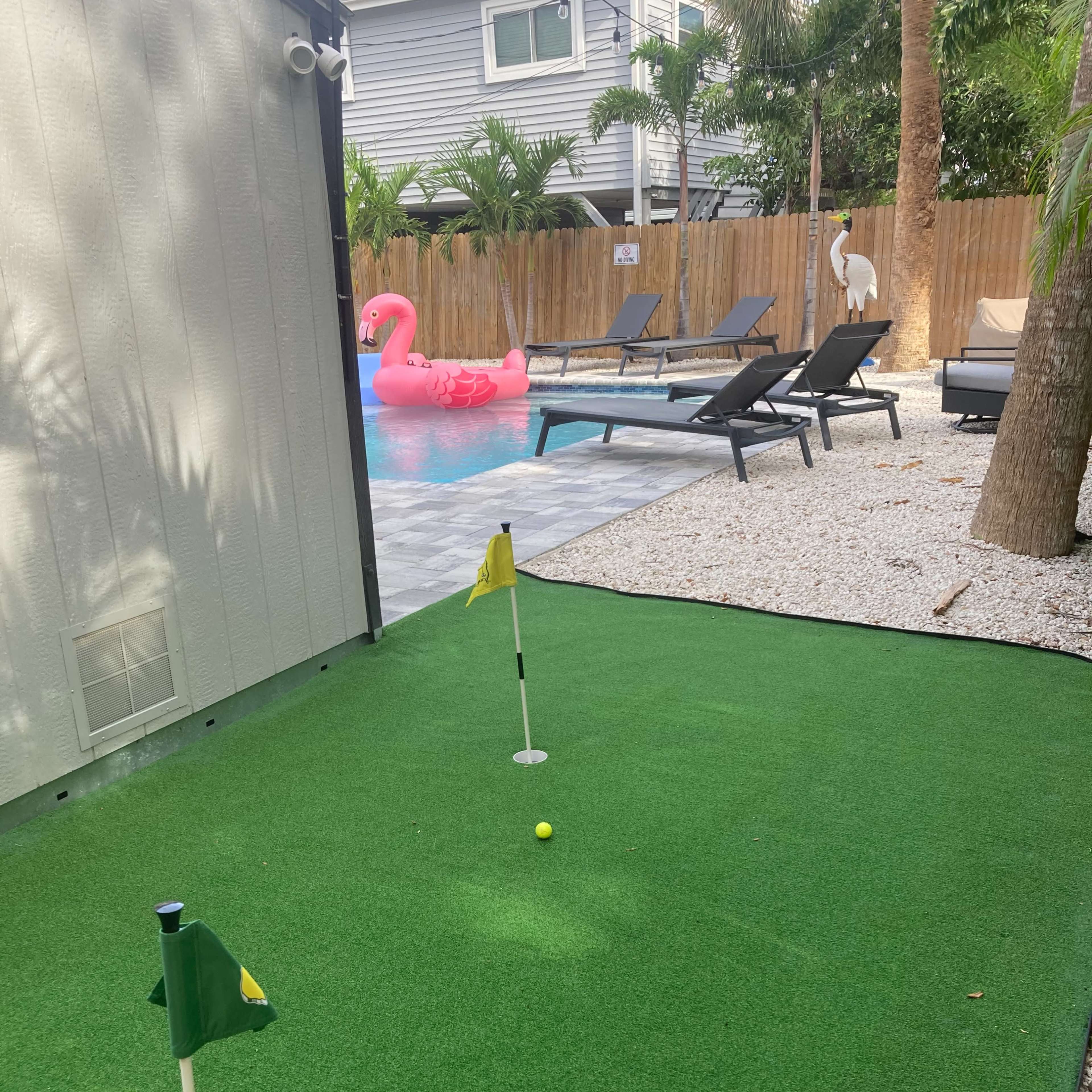 A putting green with two flags in a backyard featuring a swimming pool, lounge chairs, and a pink inflatable flamingo.