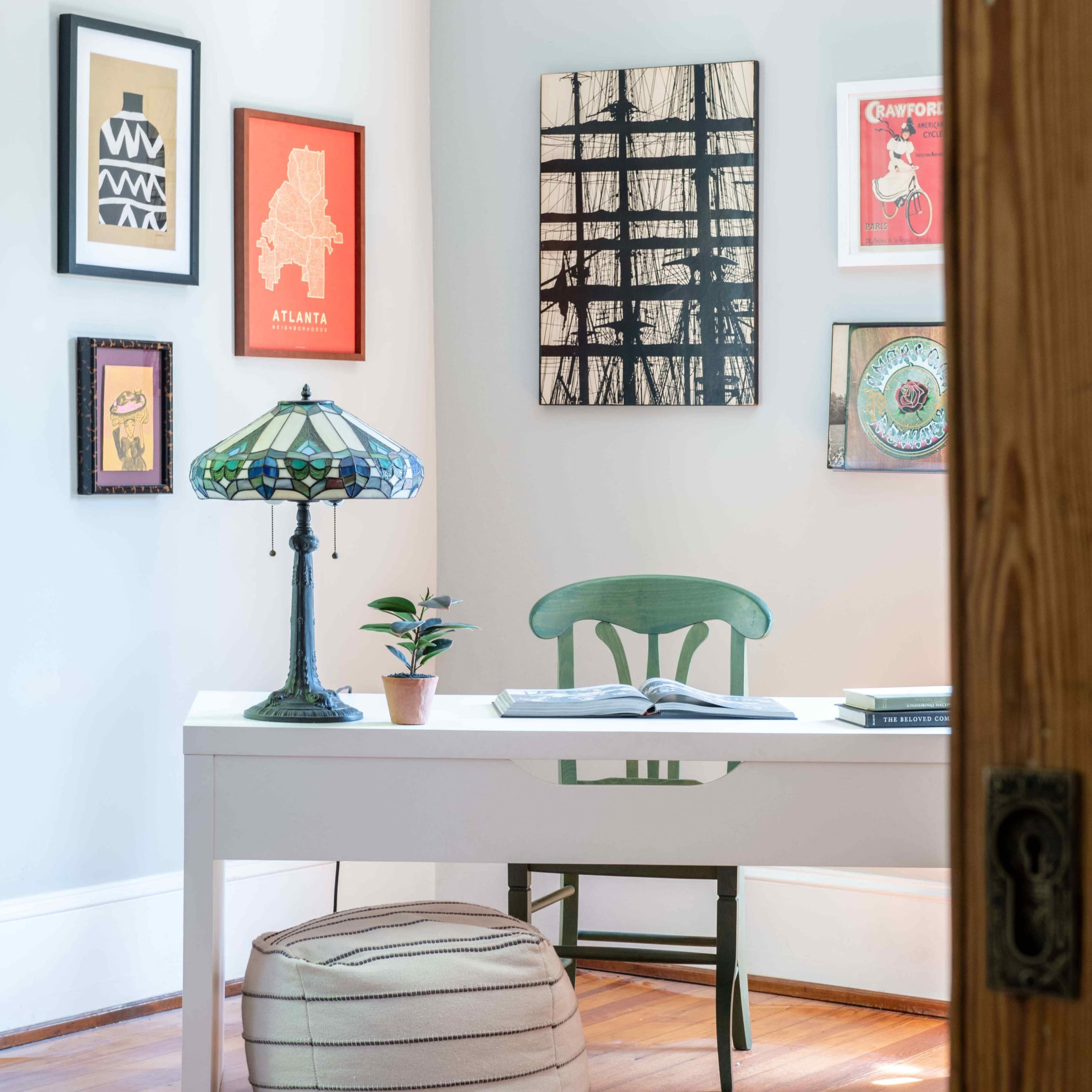 A small workspace features a white desk with an open book, a green chair, a potted plant, and a stained glass lamp, surrounded by framed artwork on the walls.