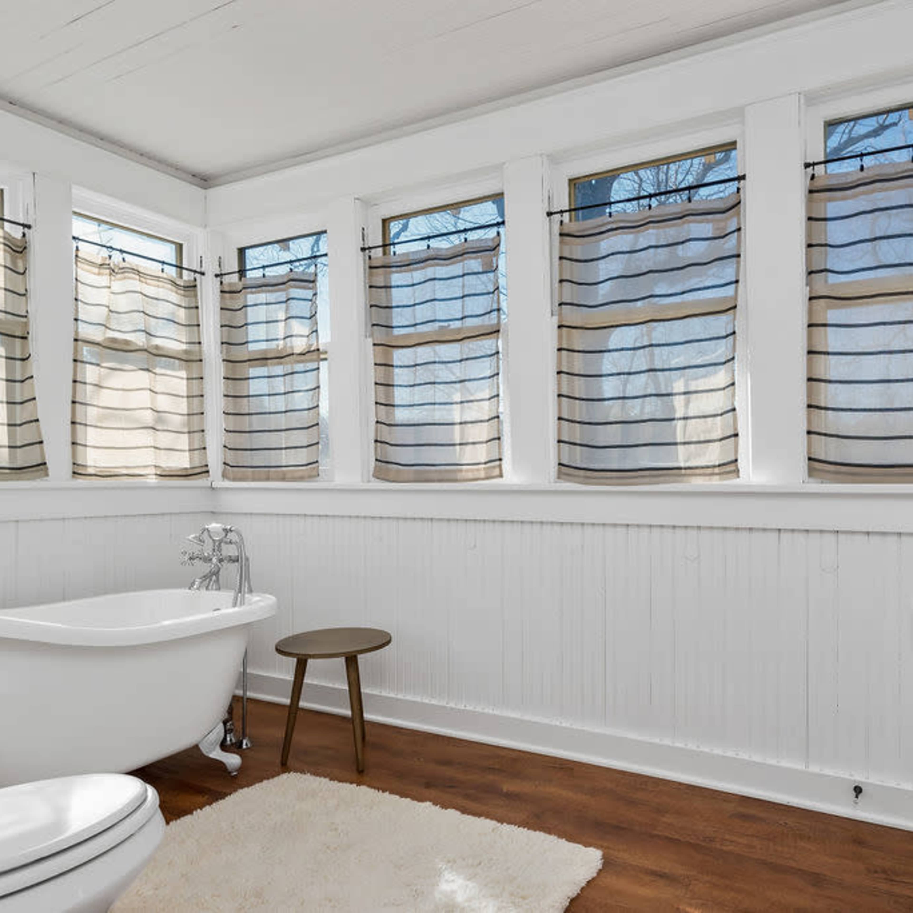 A bathroom with a freestanding bathtub, a small wooden stool, and a sink, featuring large windows covered with light curtains.