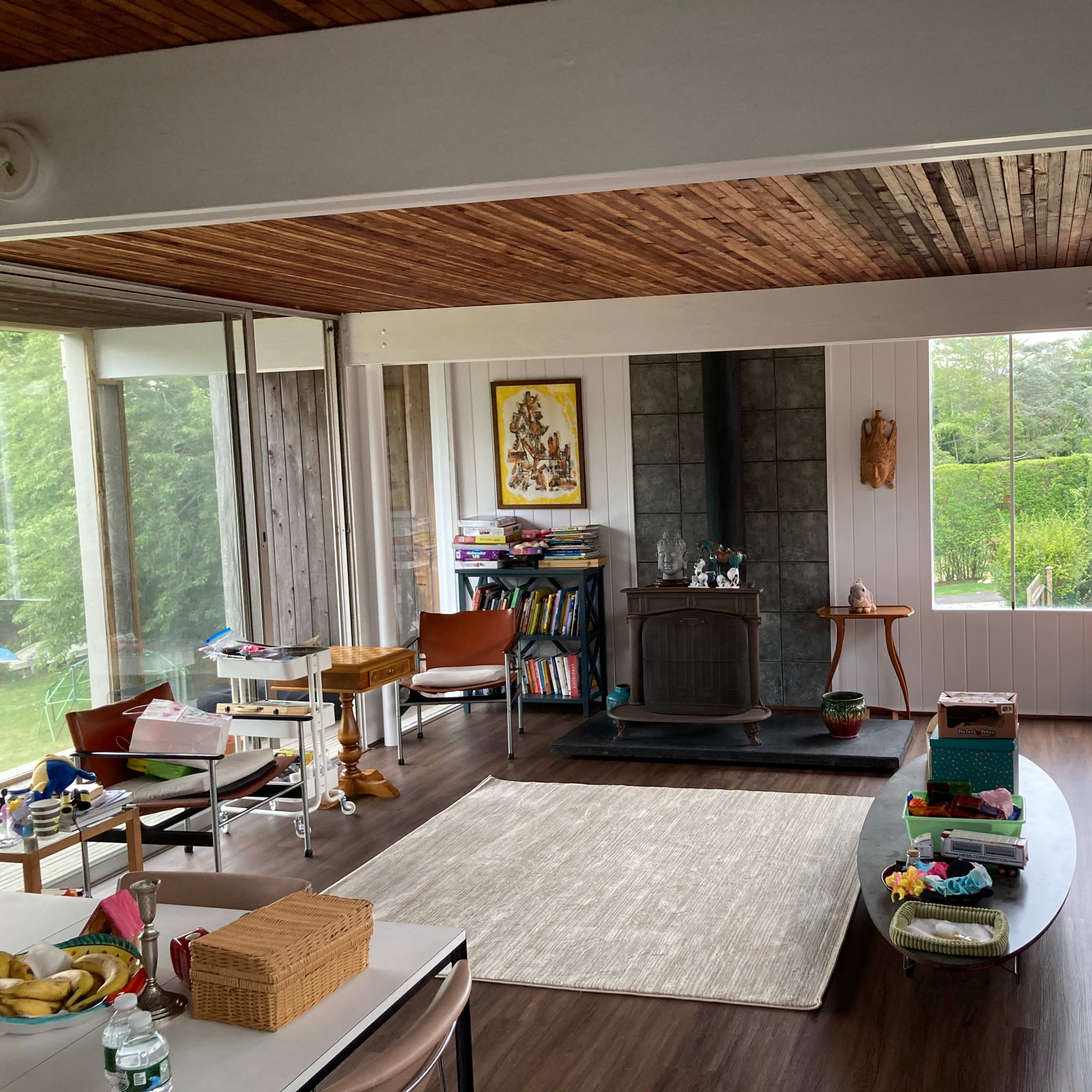 A spacious living room with wooden ceilings, a central fireplace, and large windows overlooking a green outdoor area, furnished with a red sofa and various items on tables.