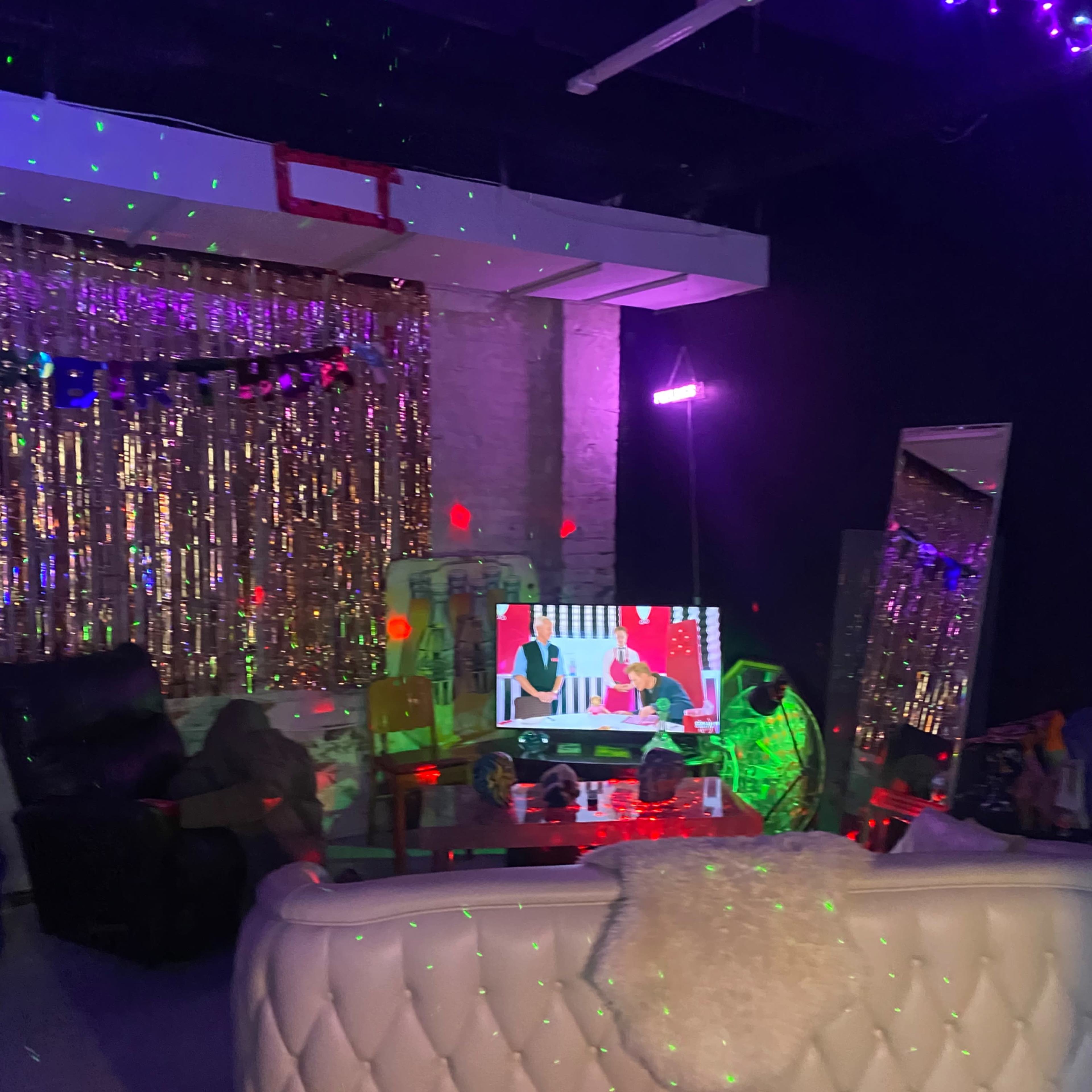 A dimly lit room with a white couch, a television displaying a show, decorated walls with shiny curtains, and colorful lights casting patterns across the space.