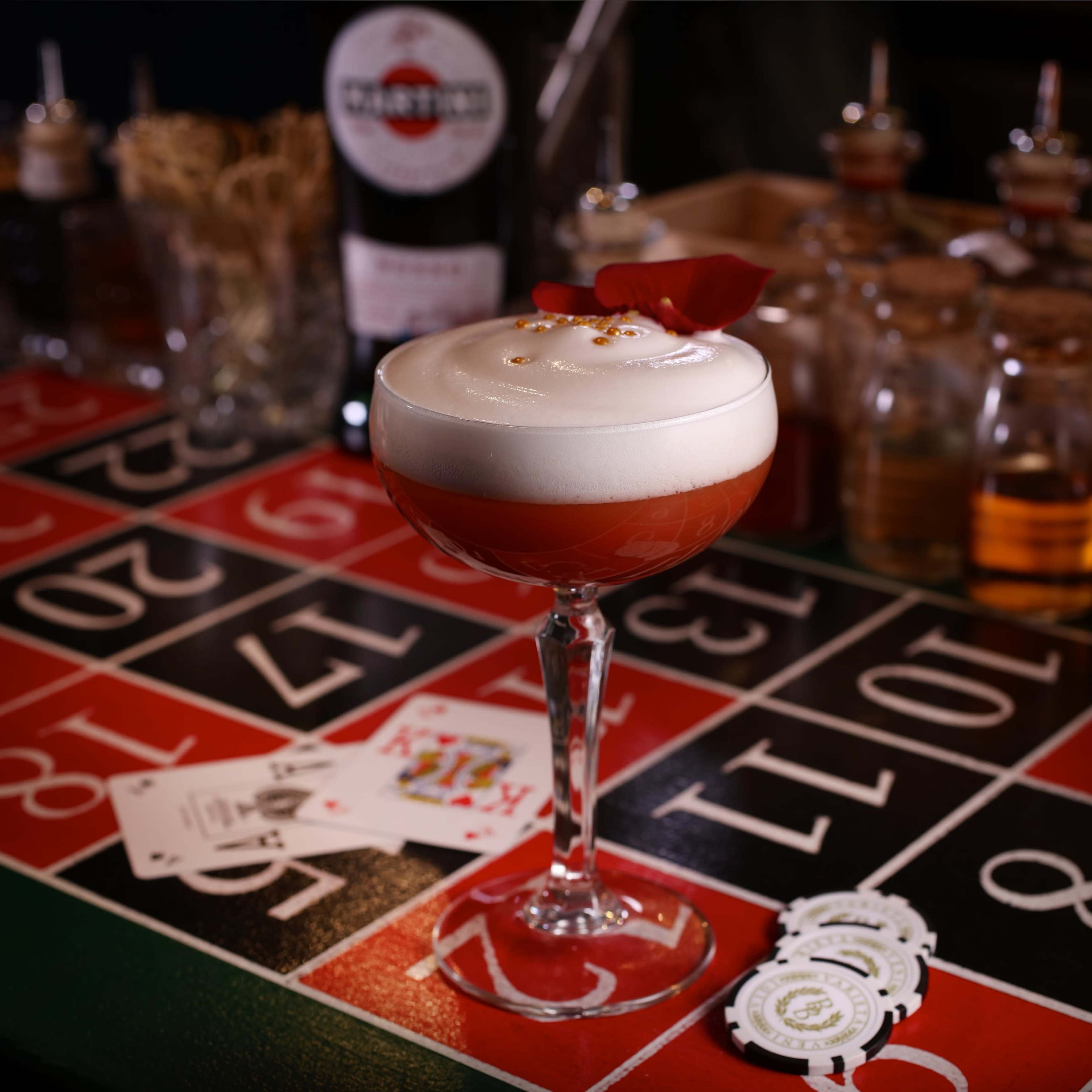 A cocktail is seated on a roulette table, surrounded by playing cards and casino chips, with various bottles and jars in the background.