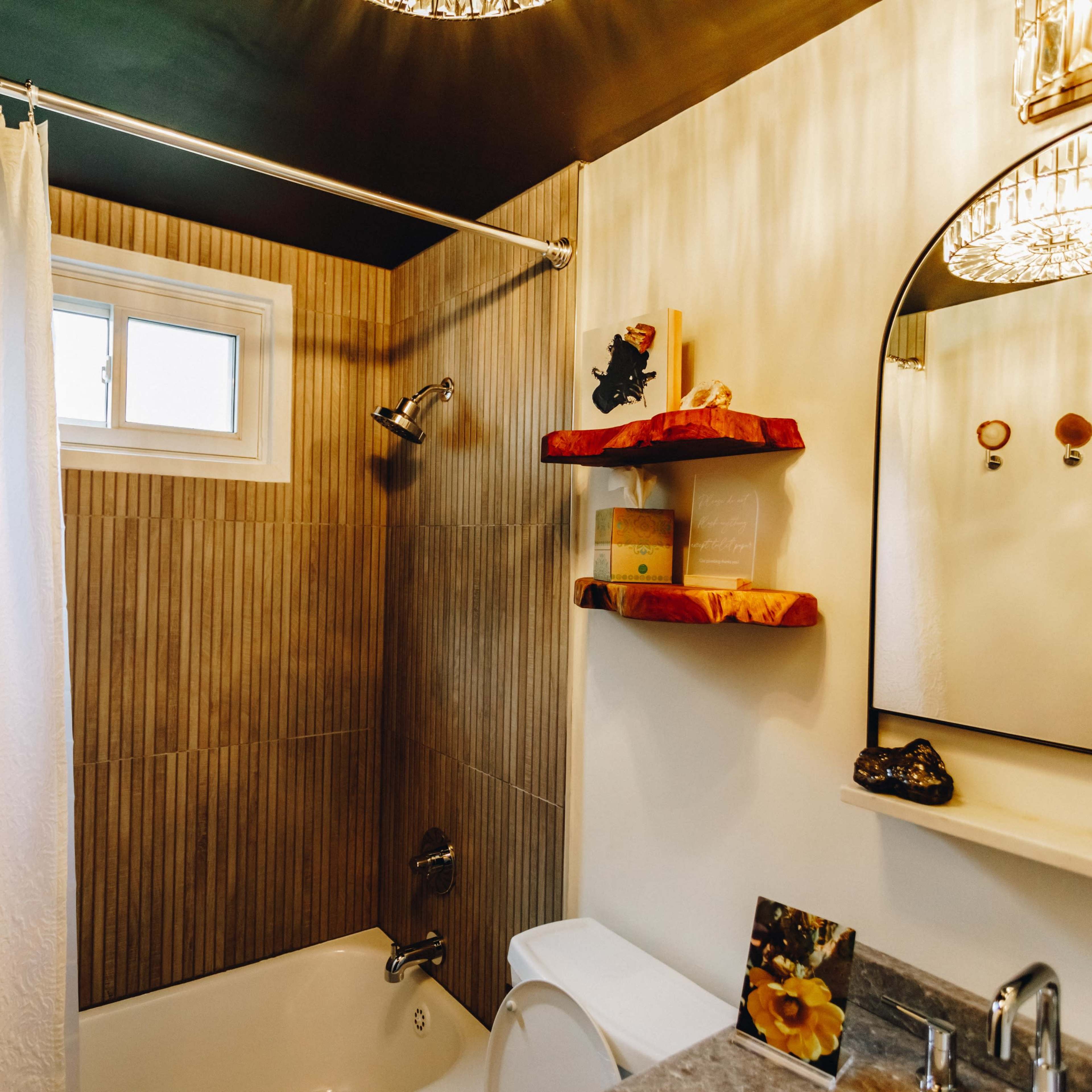 A compact bathroom features a bathtub with a shower, a window, decorative shelves, and a round mirror with two light fixtures above.