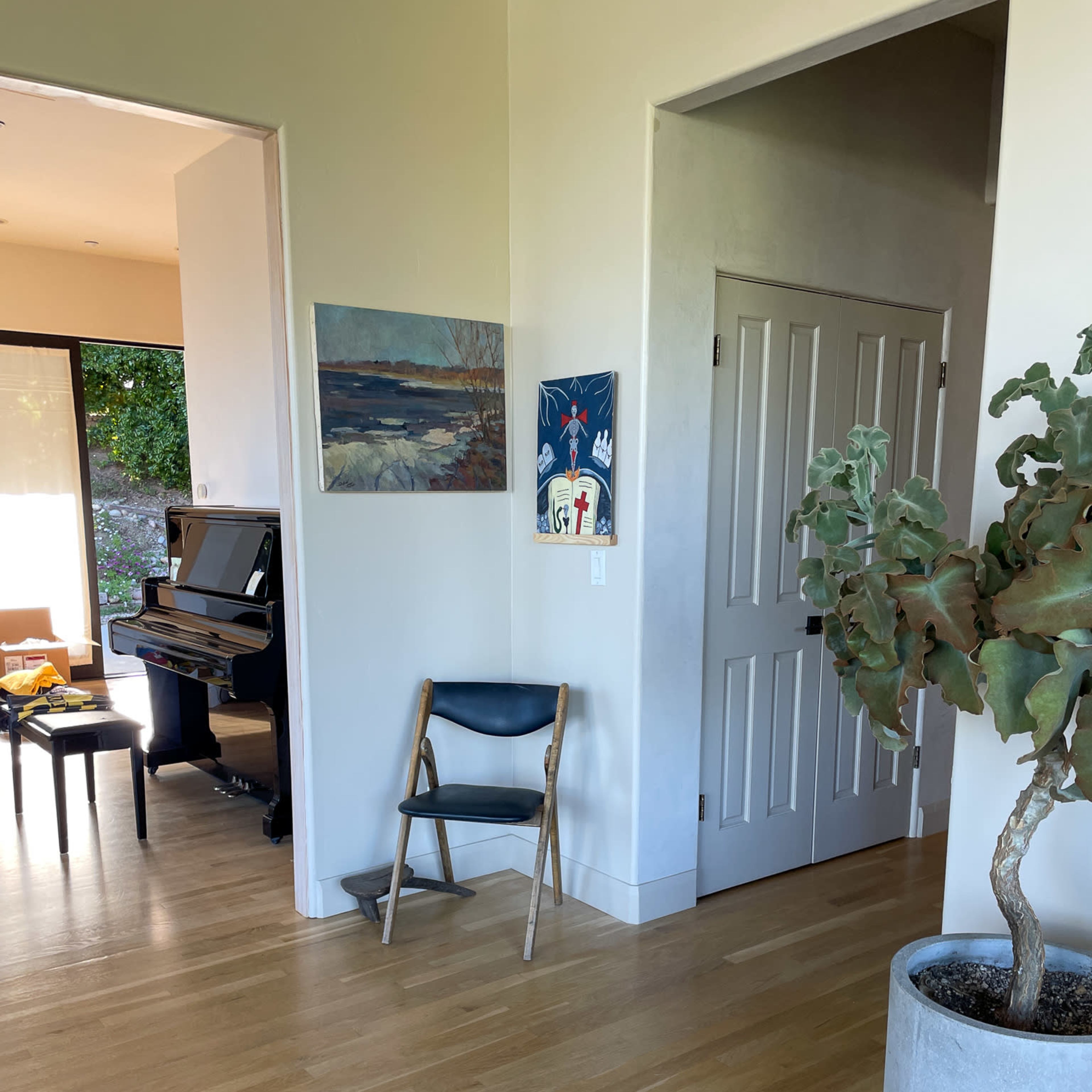 A bright, open hallway features a wooden floor, a small plant in a large pot, a folding chair, and a doorway leading to a room with a piano.