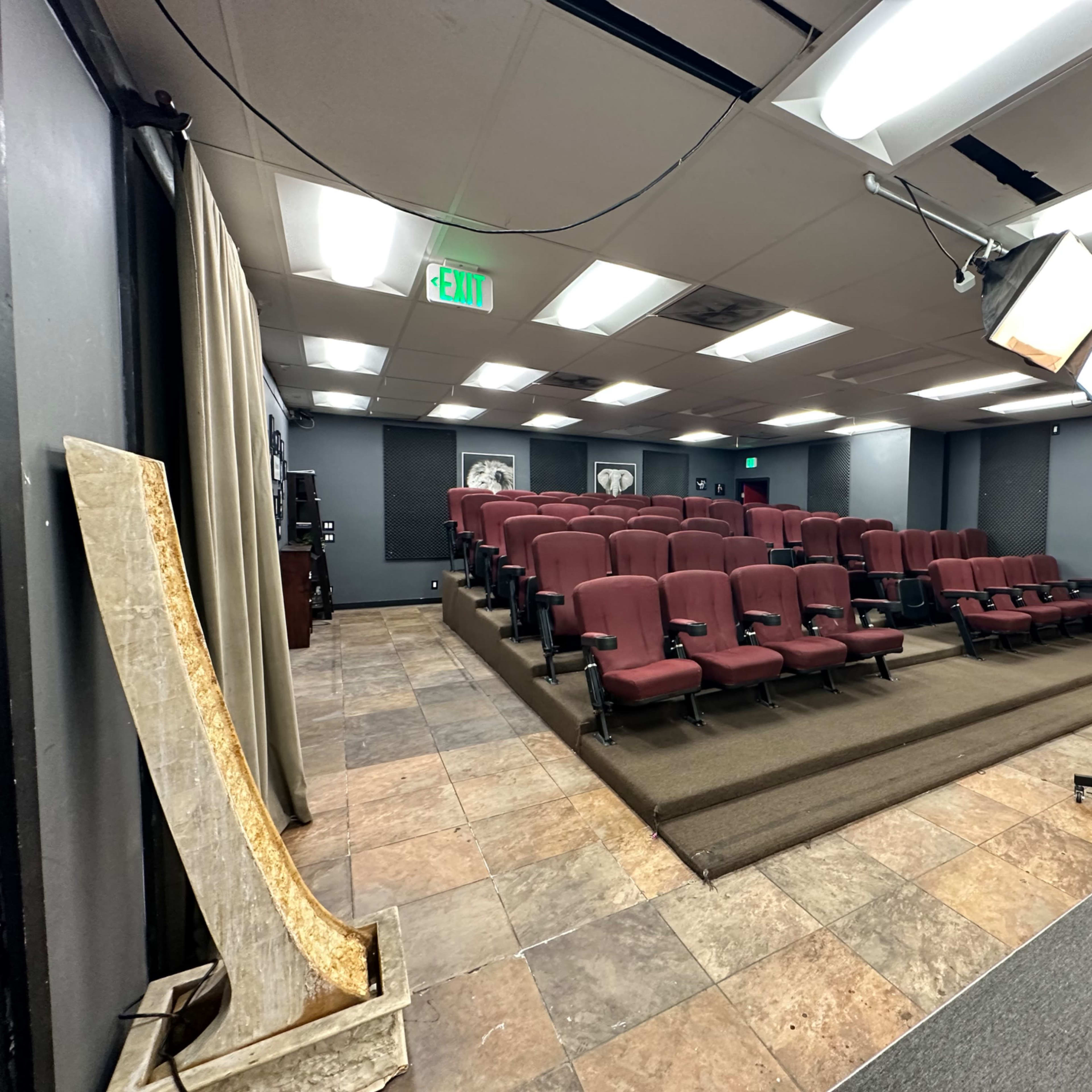 A small auditorium with rows of burgundy seating, a stage setup with lighting equipment, and a tiled floor.