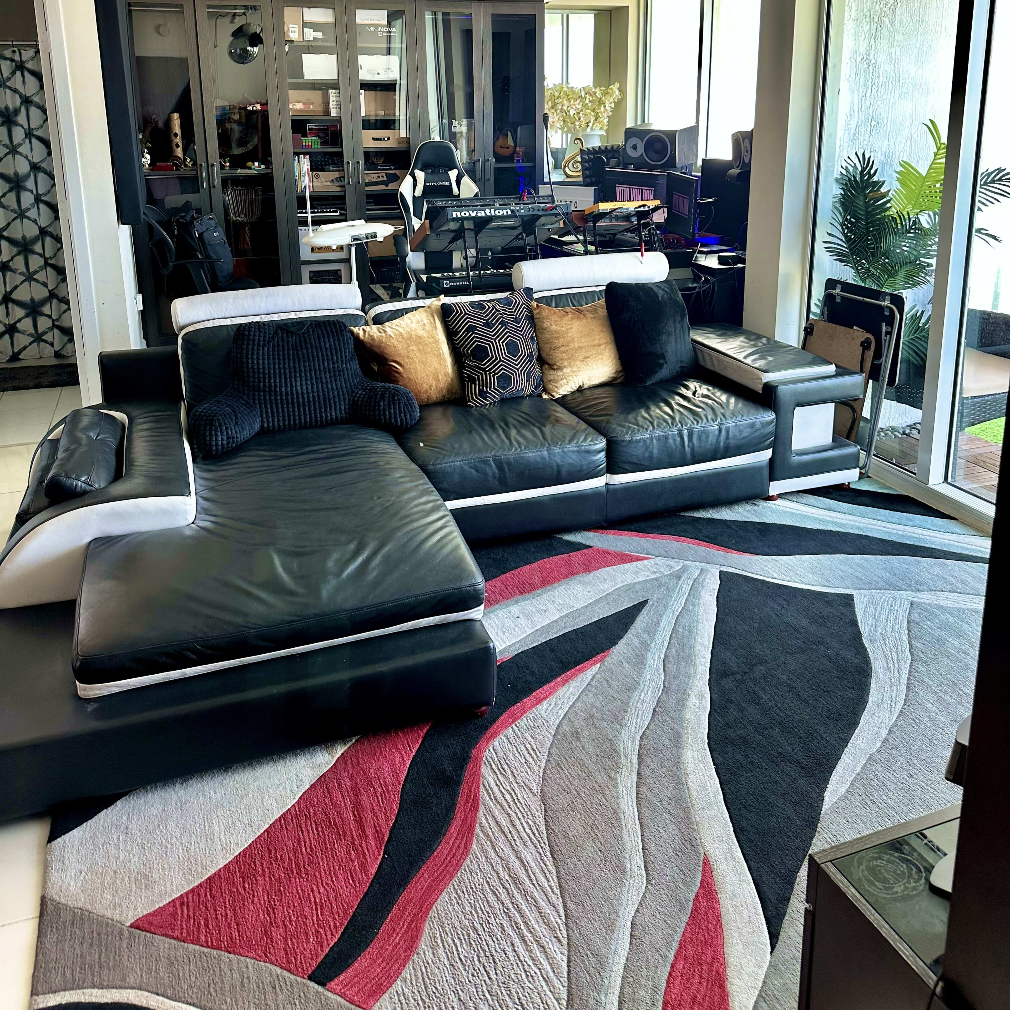 A modern living room with a large black sectional sofa, a patterned area rug, and a workspace featuring music equipment in the background.