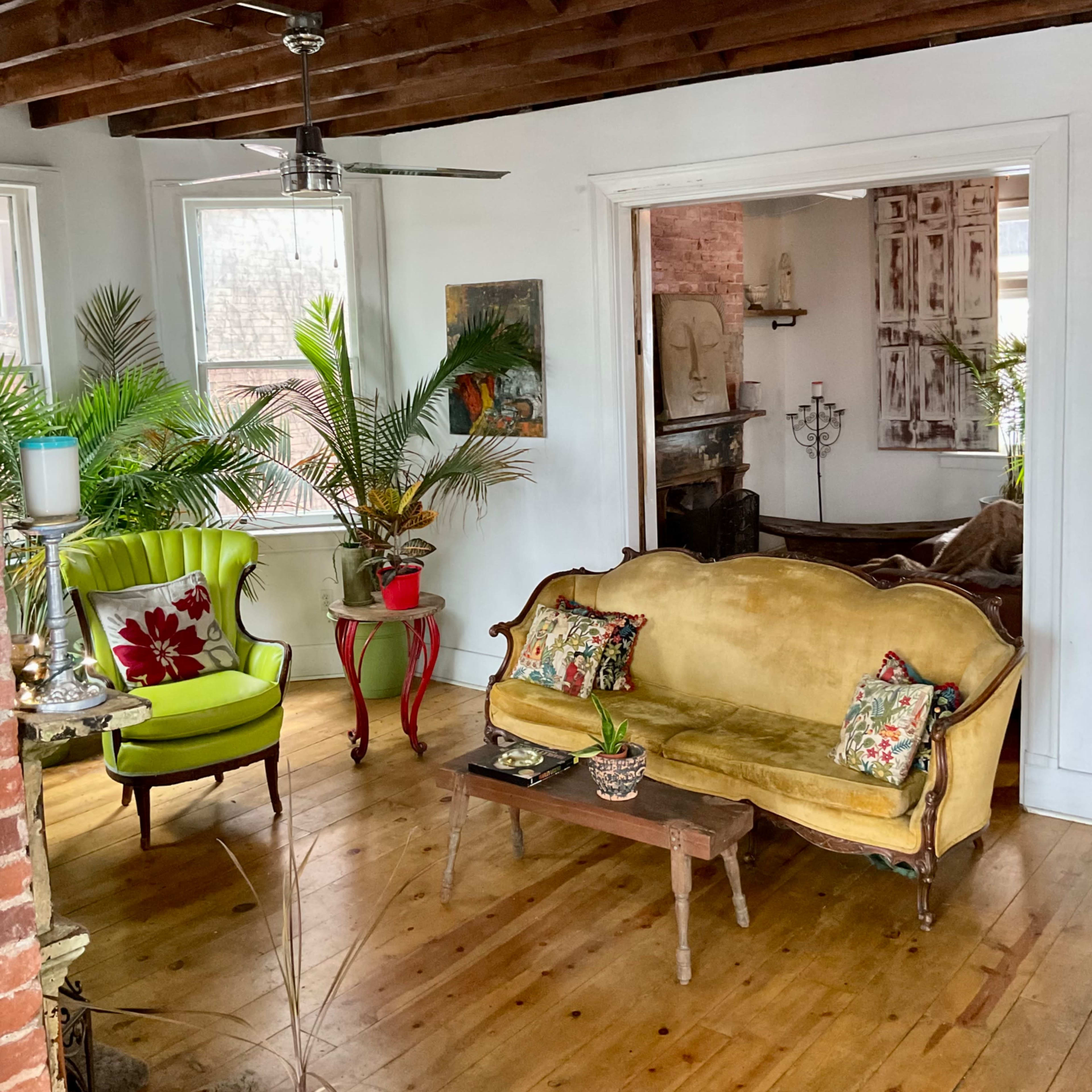 A cozy living room with a yellow sofa, a green armchair, and decorative plants, set against exposed wooden beams and brick walls.