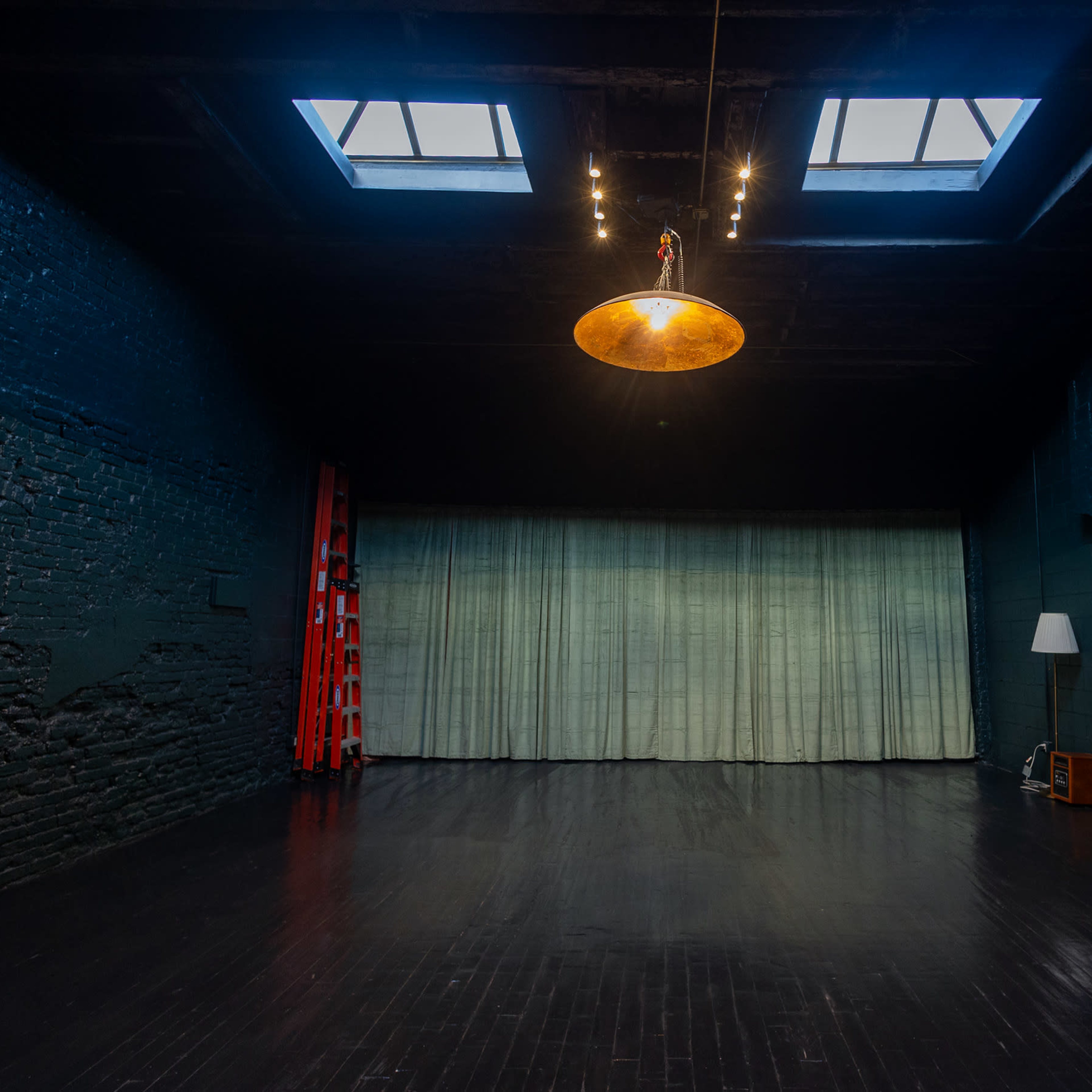 A spacious, dimly lit room with dark walls, a wooden floor, a suspended light fixture, a green curtain, a ladder, and a lamp in the corner.