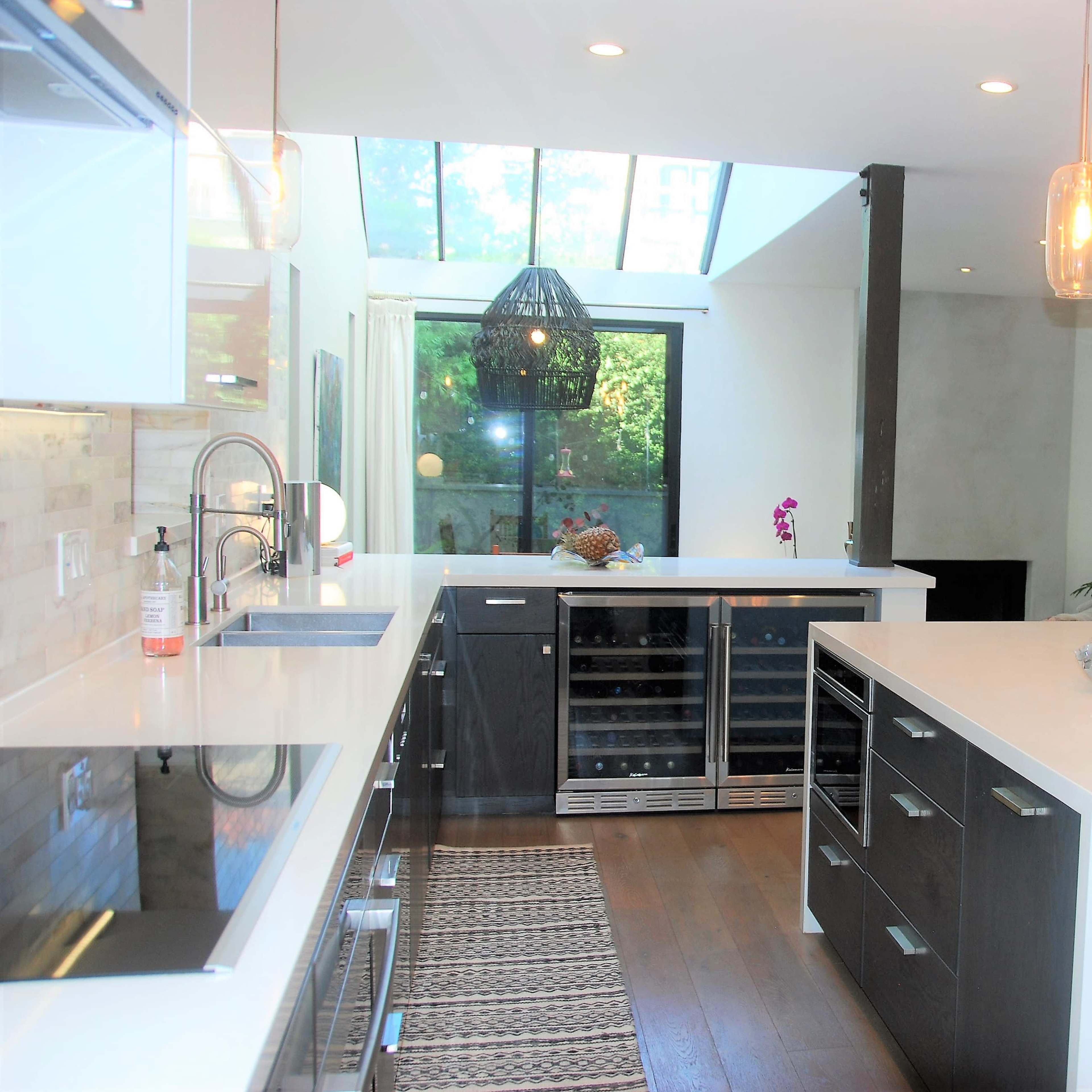 A modern kitchen features sleek cabinets, a large island with a white countertop, and pendant lighting, next to a spacious living area with large windows.