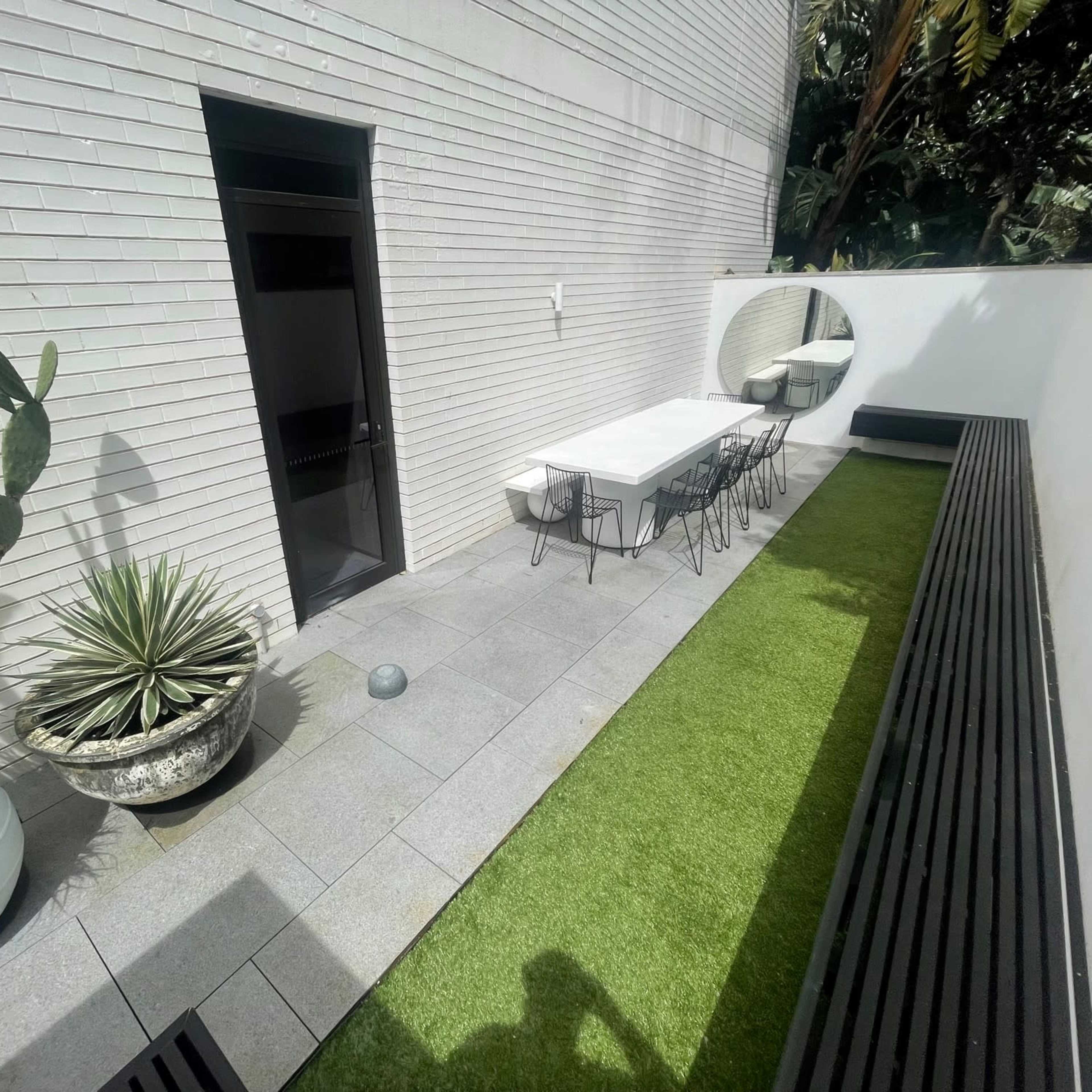 A modern outdoor space featuring a rectangular table with chairs, potted plants, and a wall with an arched mirror, all set against a backdrop of greenery.