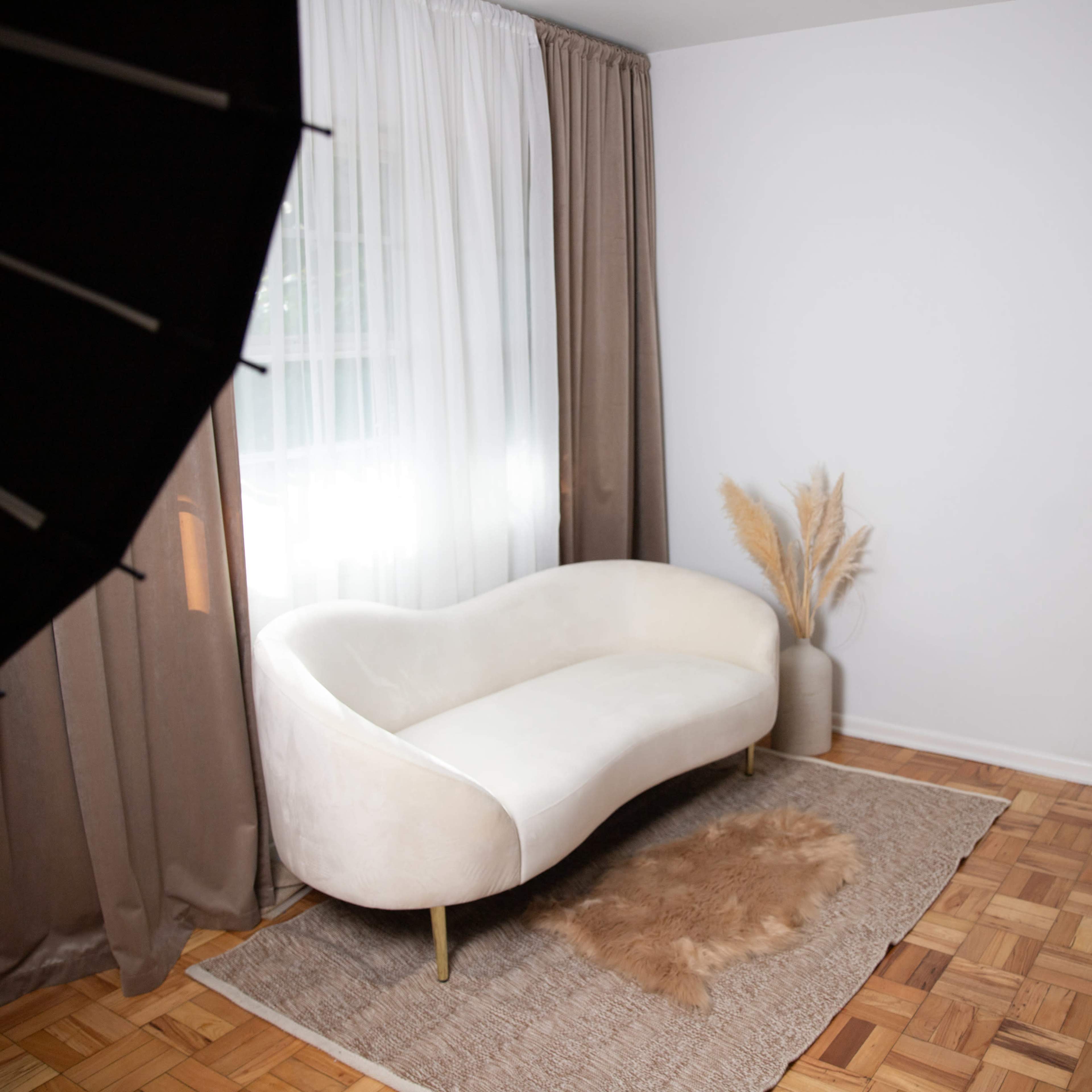 A minimalist interior with a white curved sofa, a textured rug, and decorative plants near a window with sheer curtains.