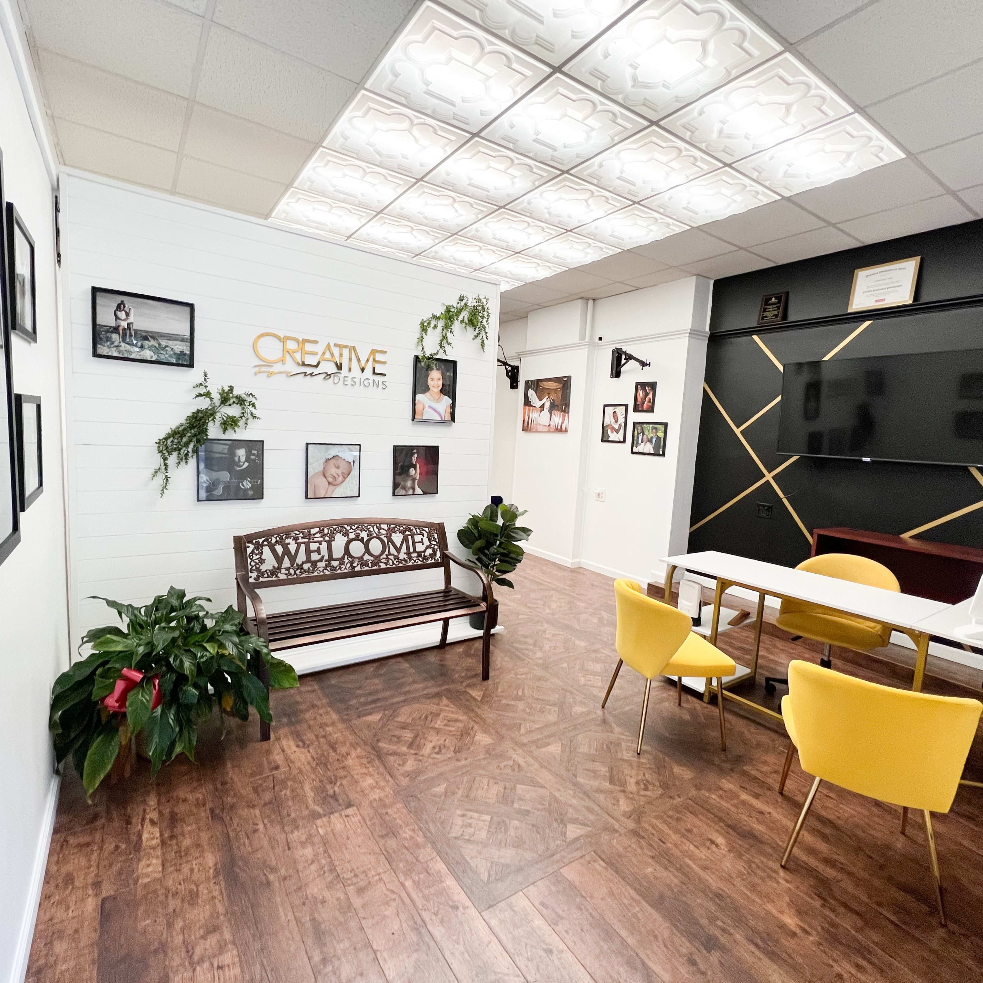 A modern office reception area featuring a wooden bench, yellow chairs, a coffee table, plants, and framed photographs on the walls.