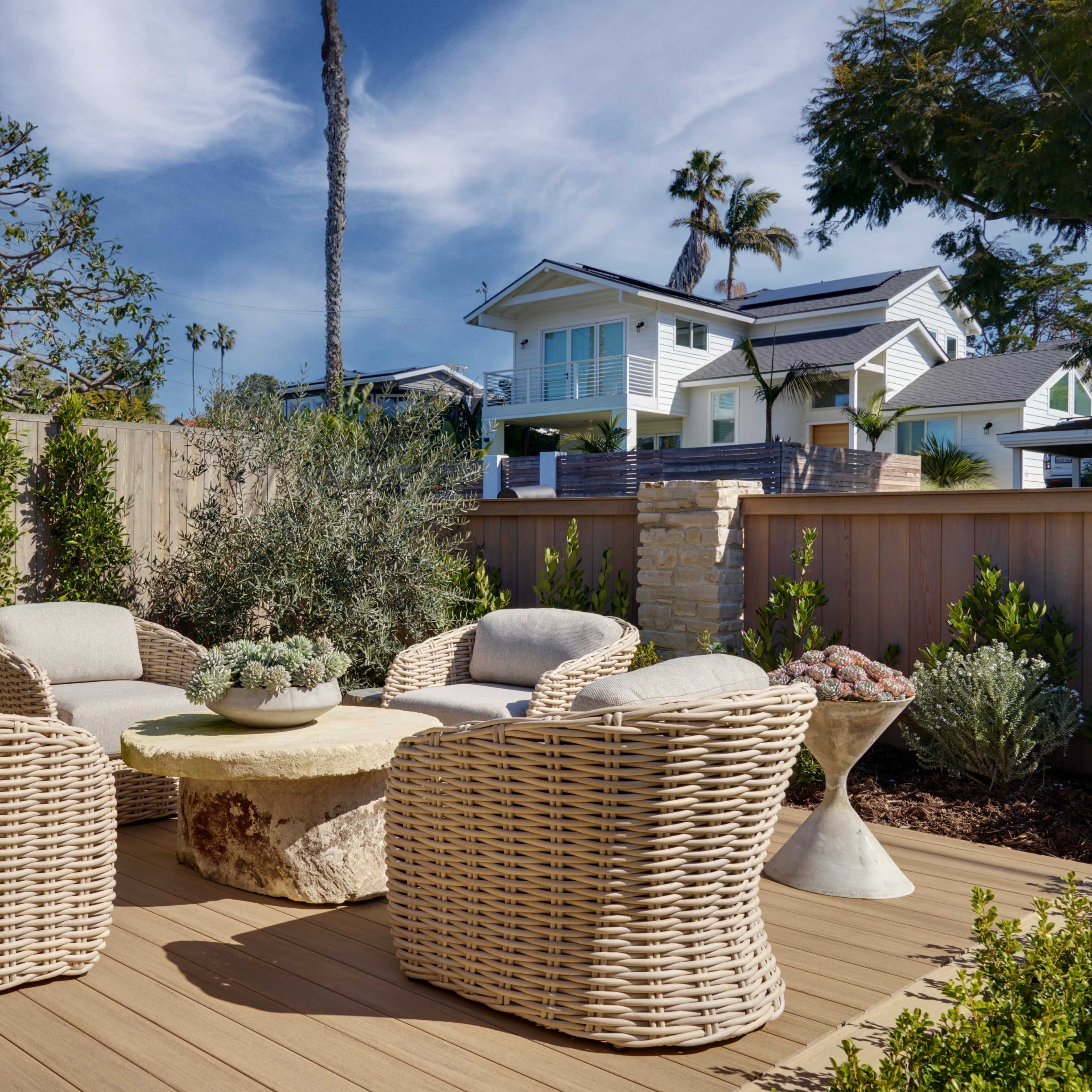 A seating area with wicker chairs and a stone table is set on a wooden deck surrounded by lush greenery and a wooden fence.