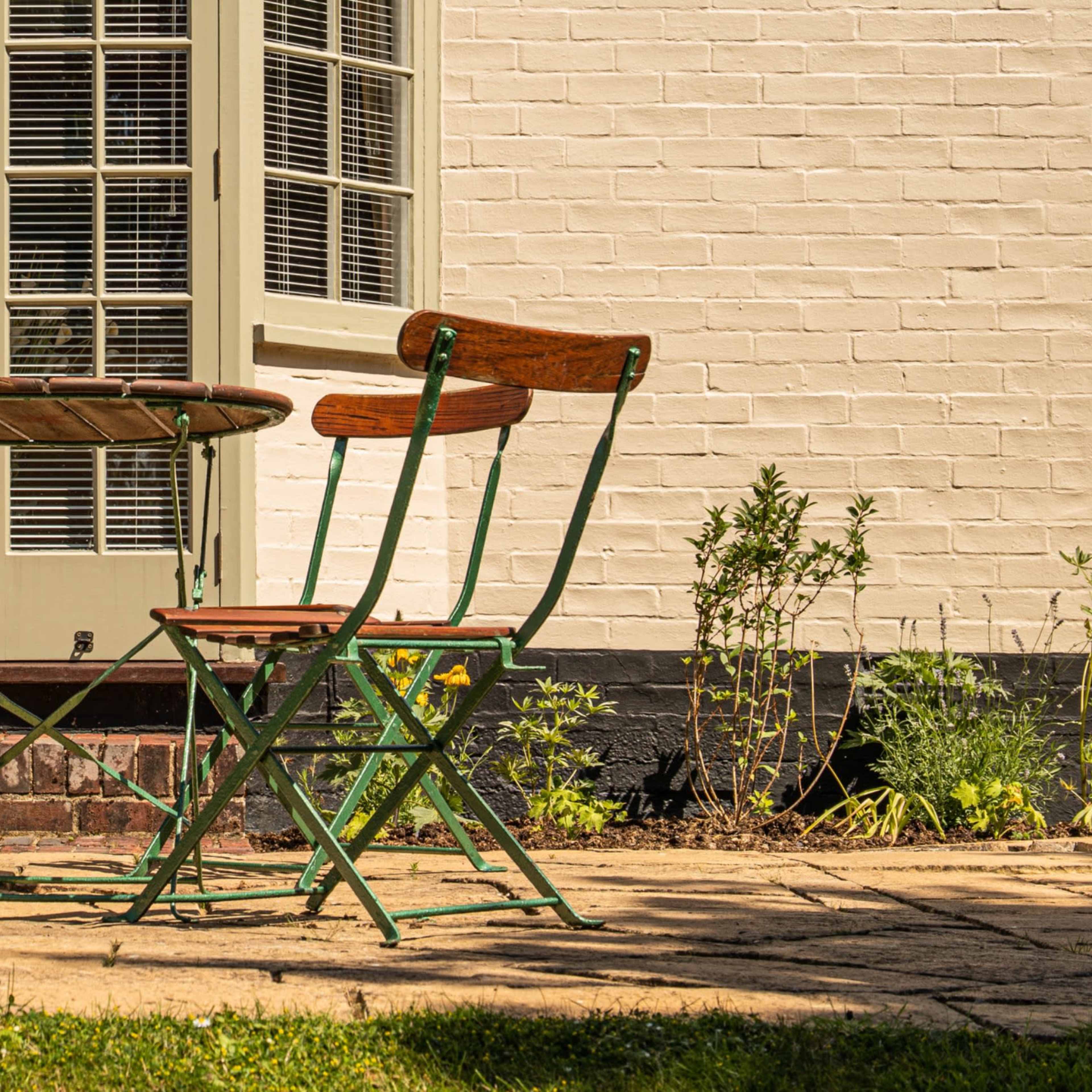 A small round table with two folding chairs sits on a stone patio beside a house with a light-colored brick wall and large windows.