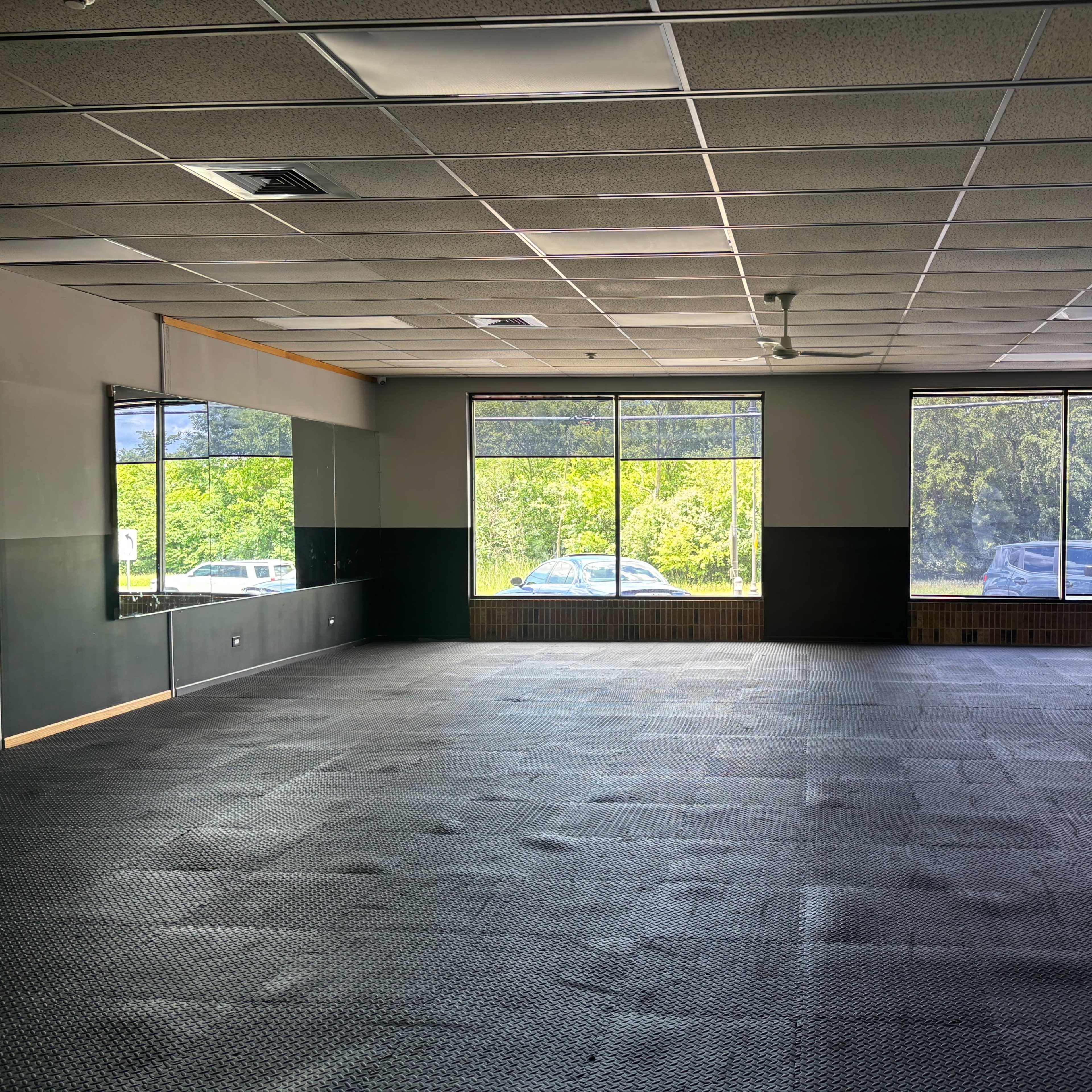 An empty, spacious room with black rubber flooring, large windows allowing natural light, and mirrors on one wall.