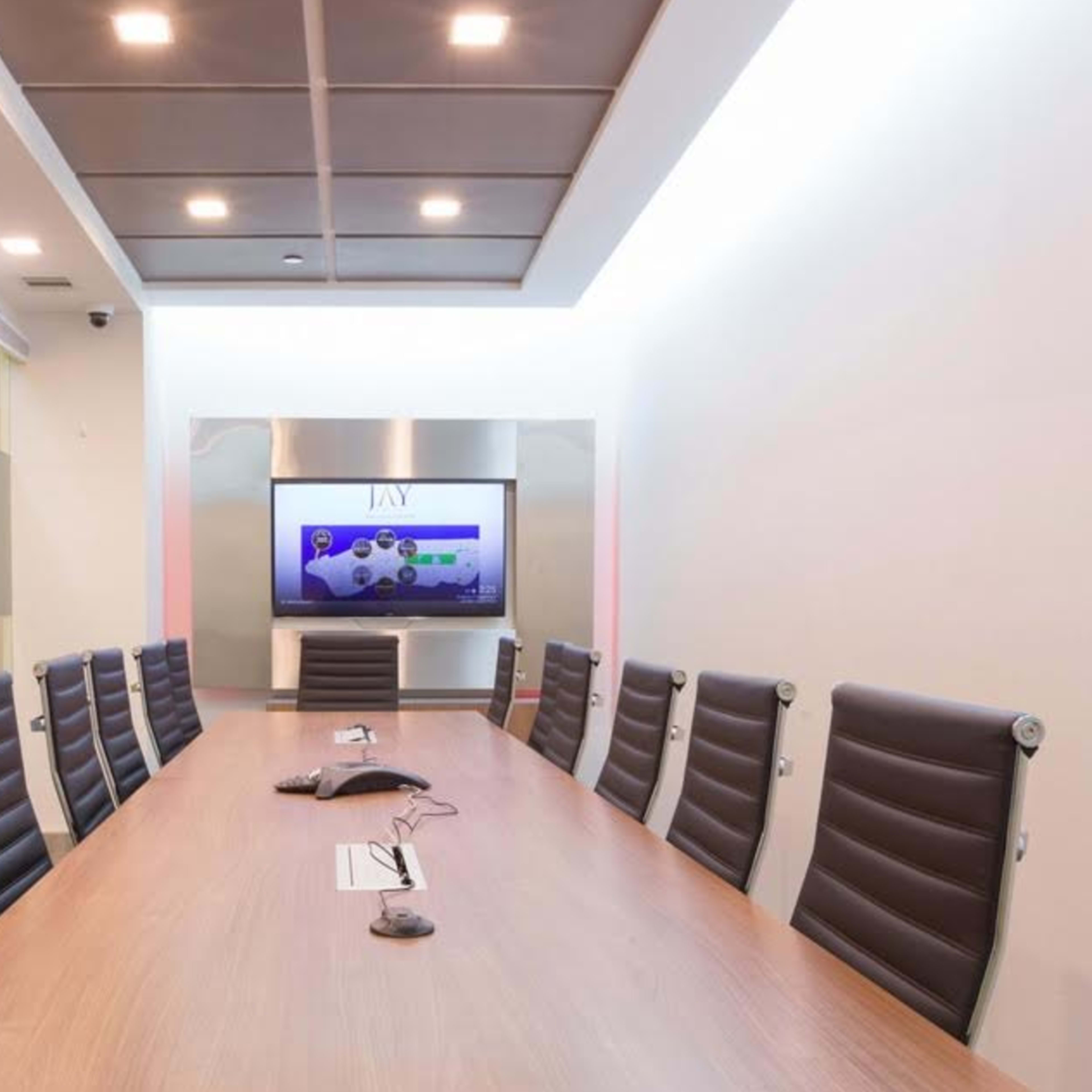 A long conference table with black leather chairs is arranged in a modern meeting room featuring a large screen at one end.