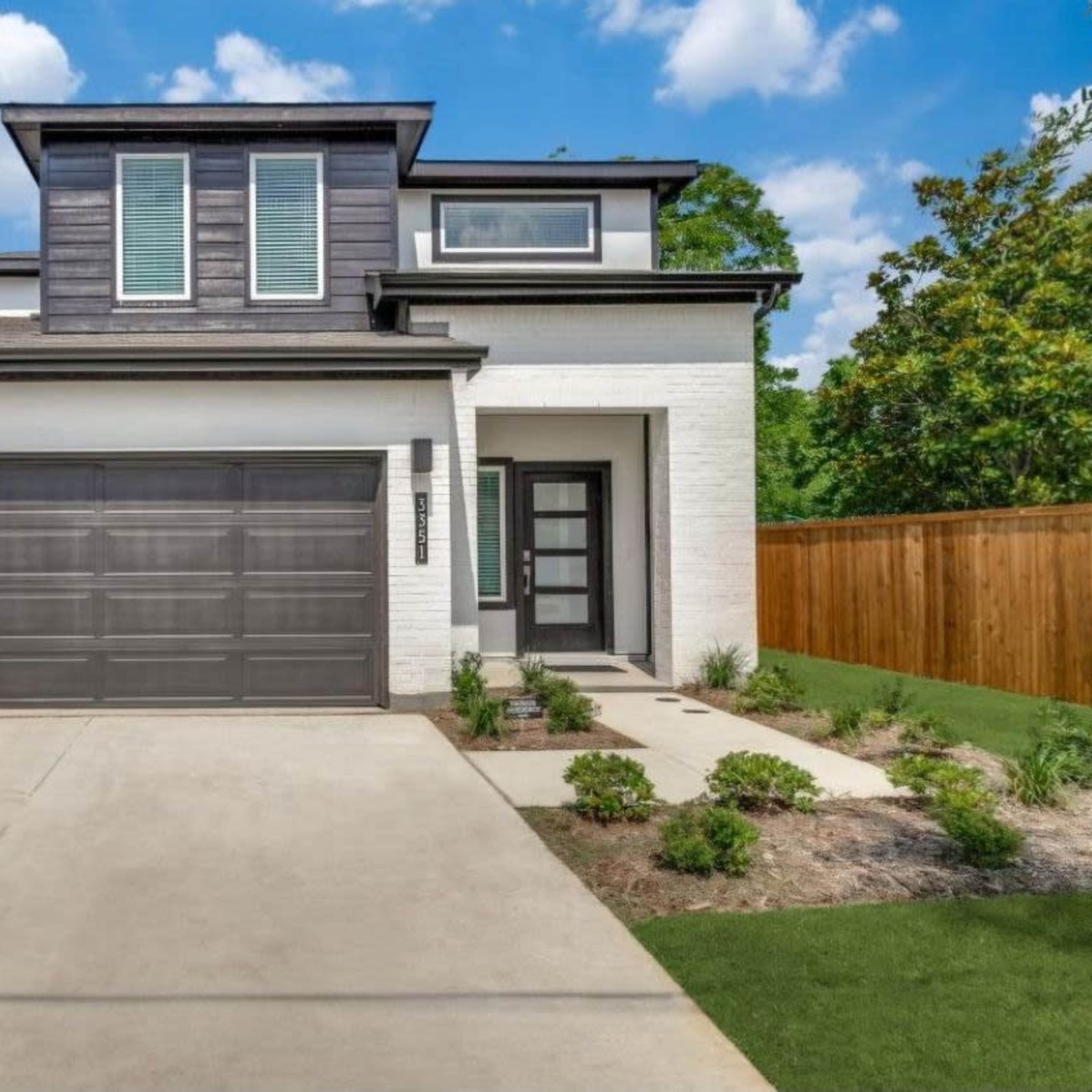 A modern two-story house with a gray garage door, a tidy front walkway, and a landscaped yard surrounded by a wooden fence.