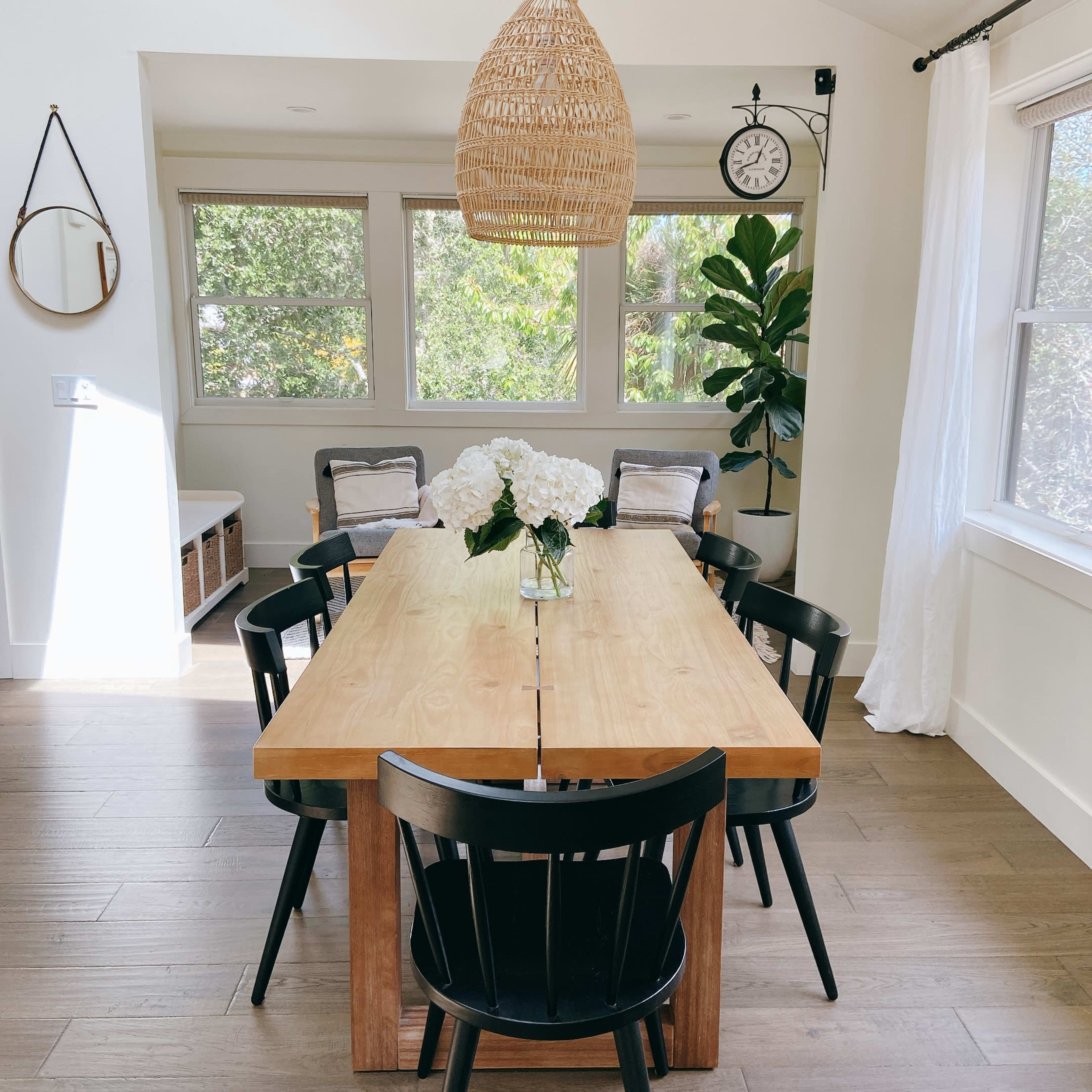 A wooden dining table with black chairs is set in a well-lit room featuring large windows and a hanging rattan light fixture.