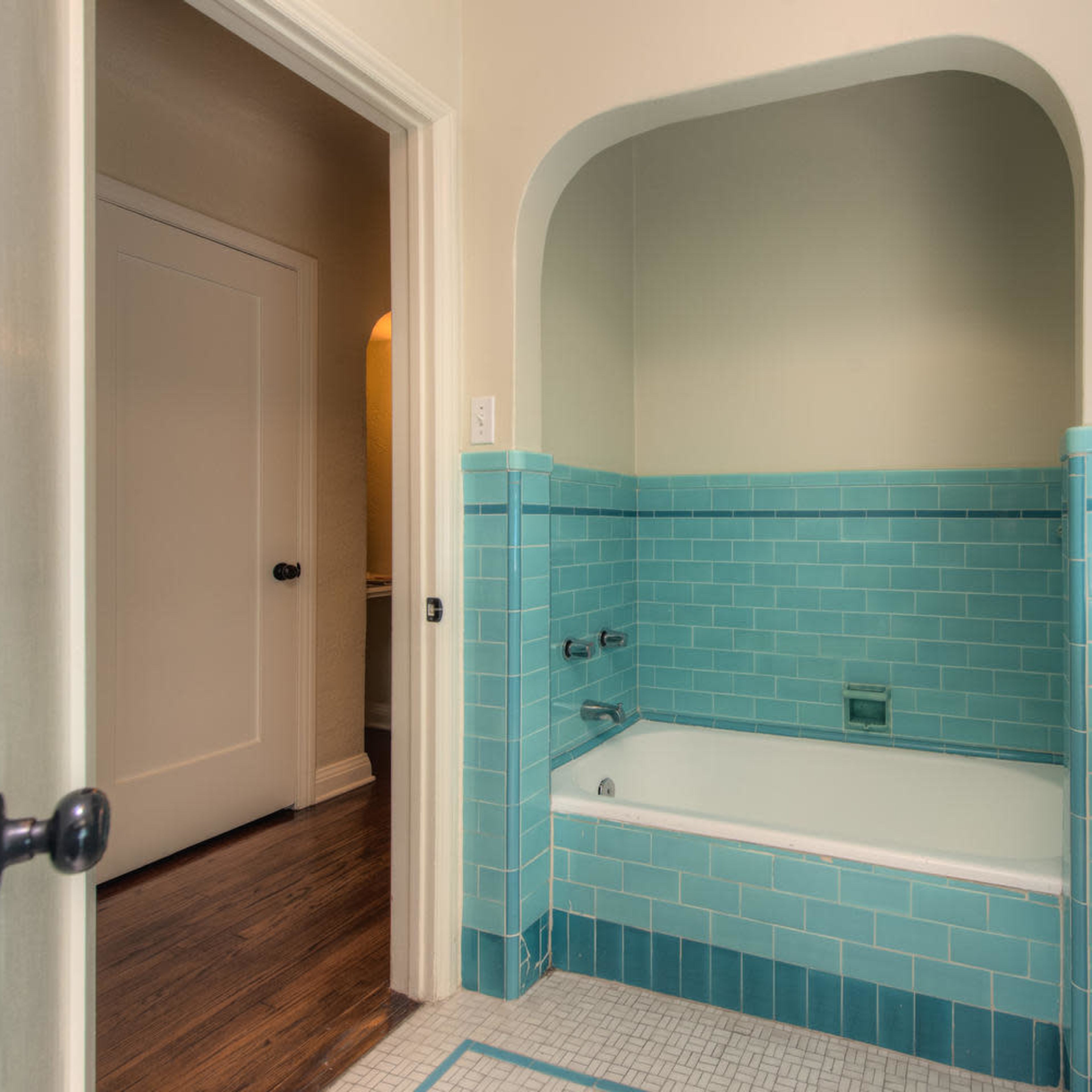 A bathroom features a turquoise tiled bathtub set within an arched alcove, with a doorway leading to another room.