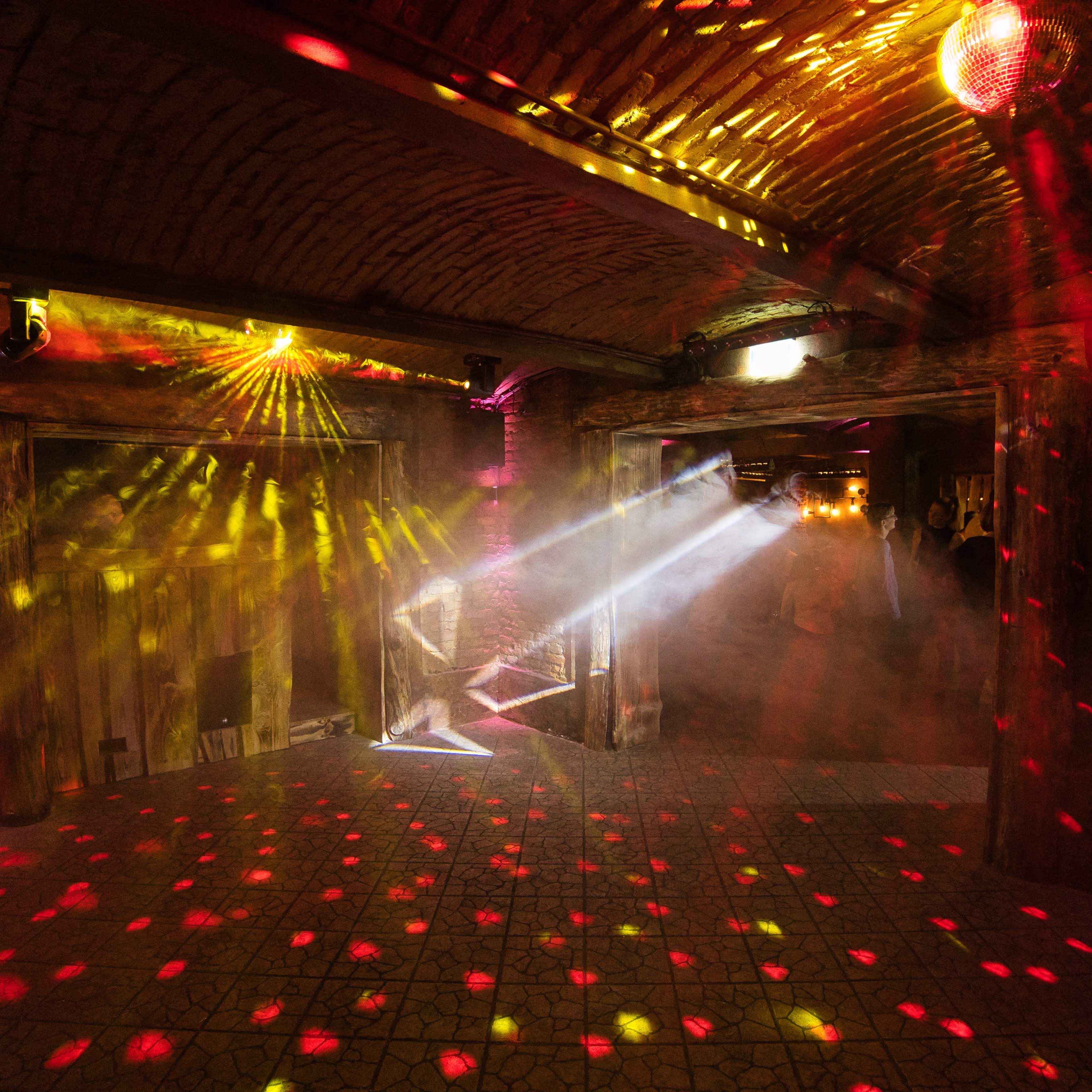 The interior of a dimly lit venue features colorful light beams and a disco ball casting patterns across the floor and walls.