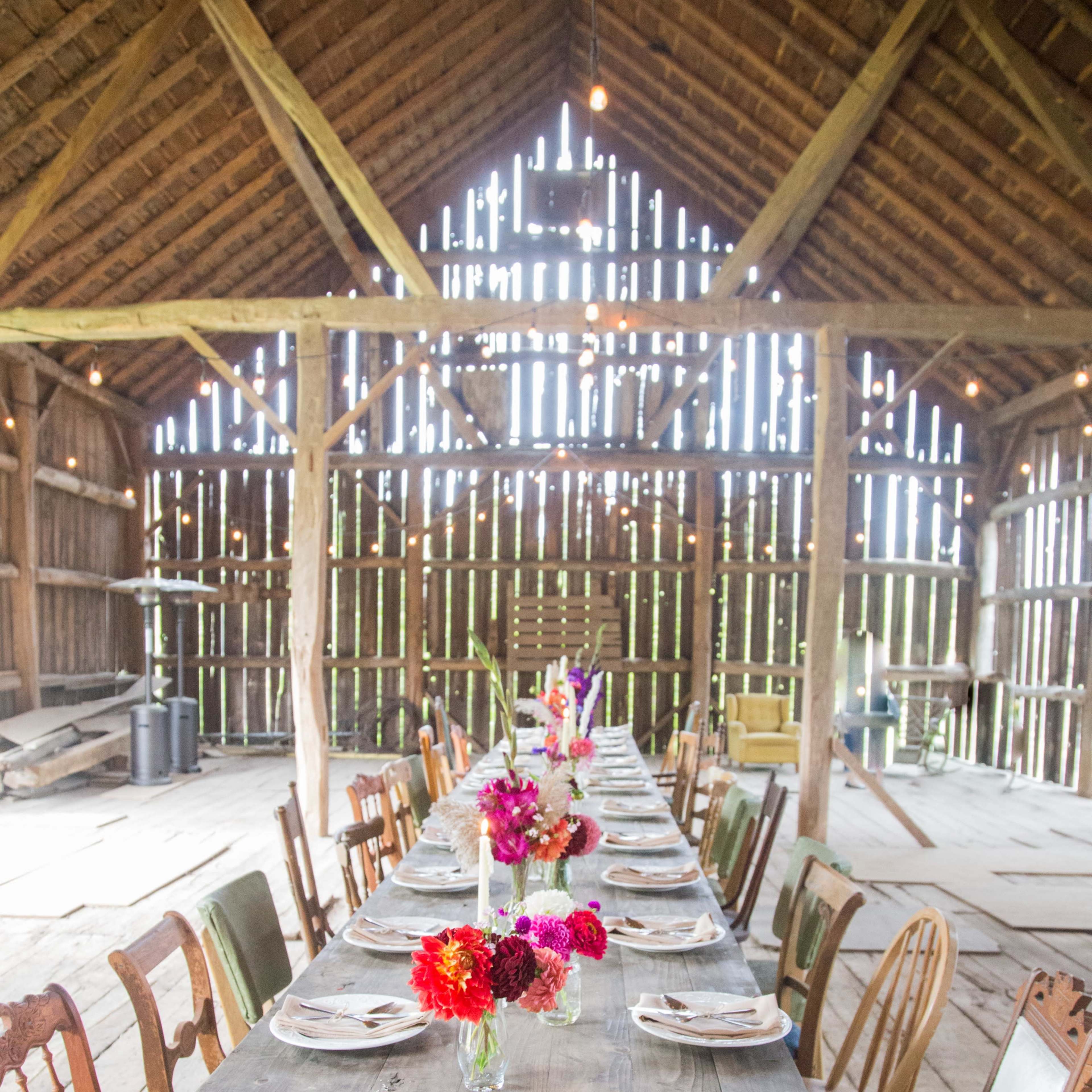 A long wooden table is set with plates and vibrant flower arrangements in a rustic barn with wooden beams and open slats.