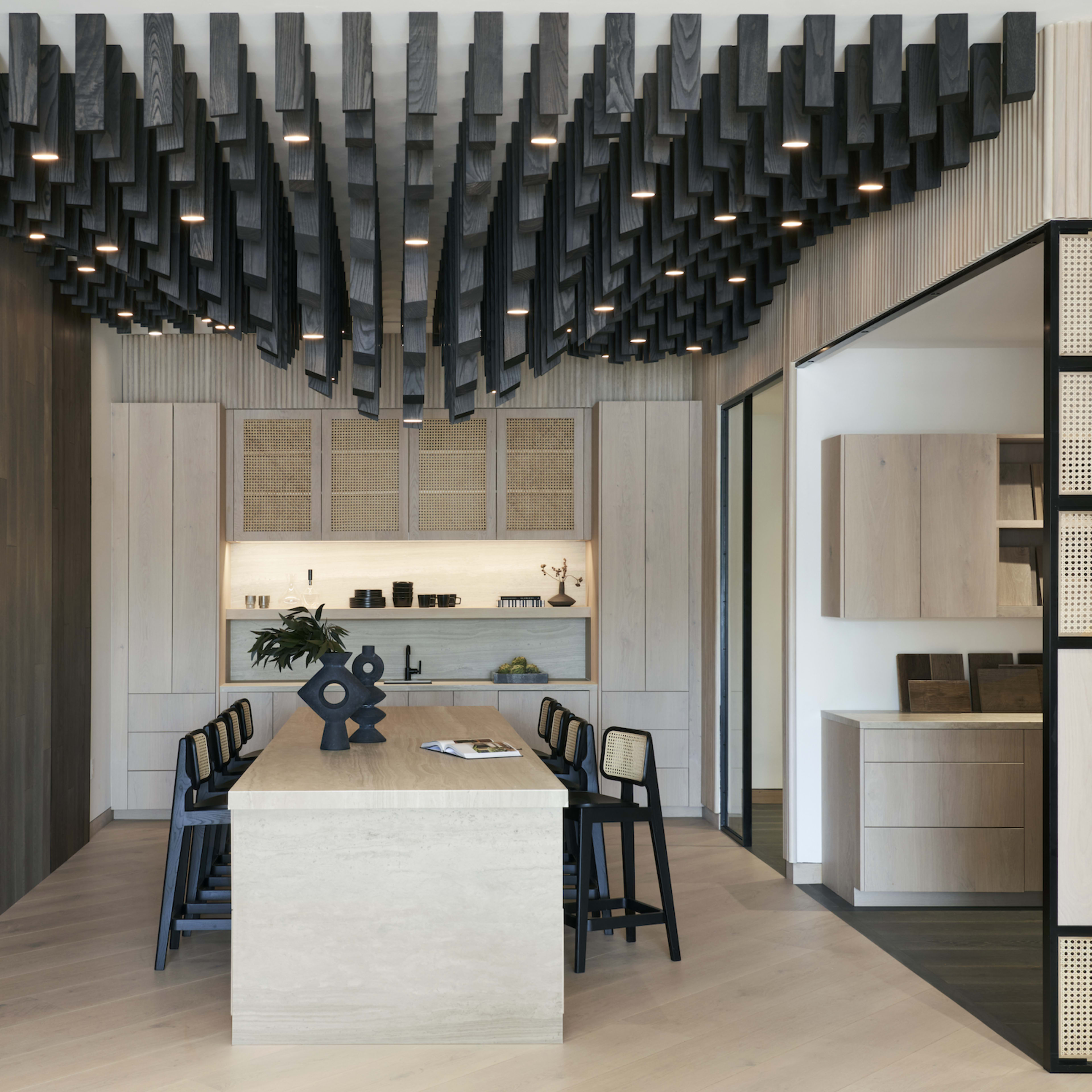 A modern dining area features a large light-colored rectangular table surrounded by black chairs, with wooden walls and a sculptural ceiling design above.