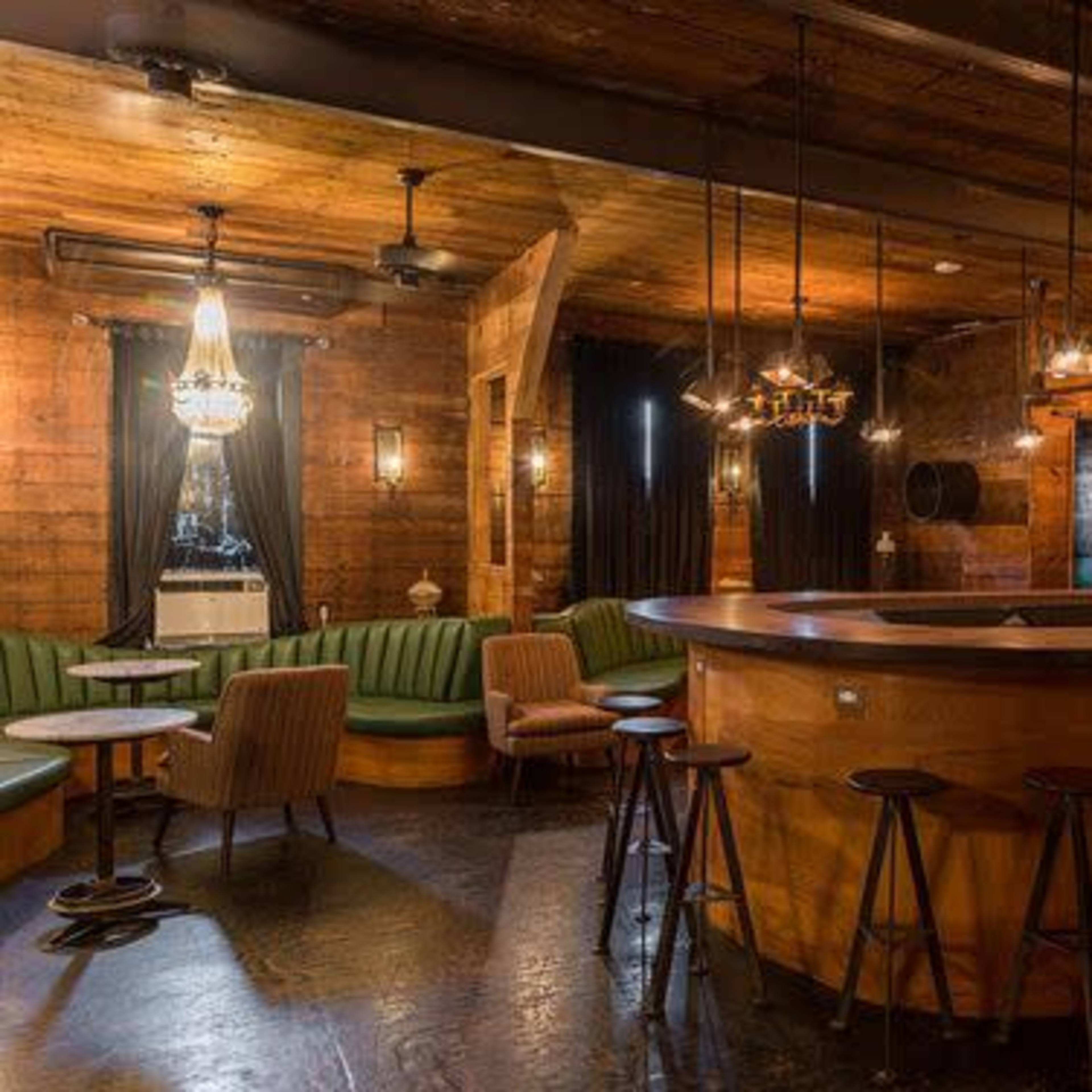 A wood-paneled bar interior with a curved wooden counter, green upholstered seating, and hanging light fixtures creating a cozy atmosphere.