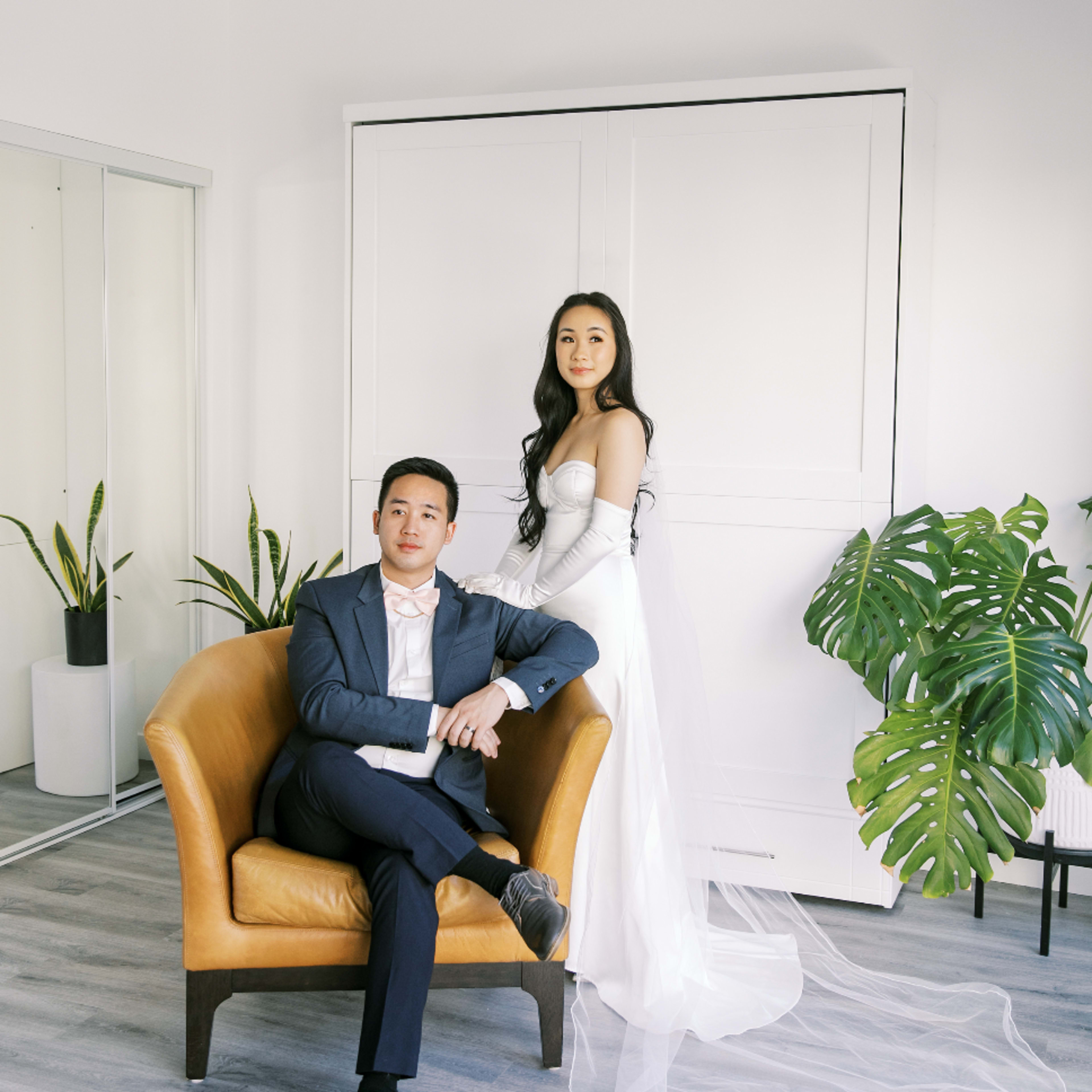 A couple poses in a modern interior, with the bride standing beside the groom who is seated on a leather armchair.