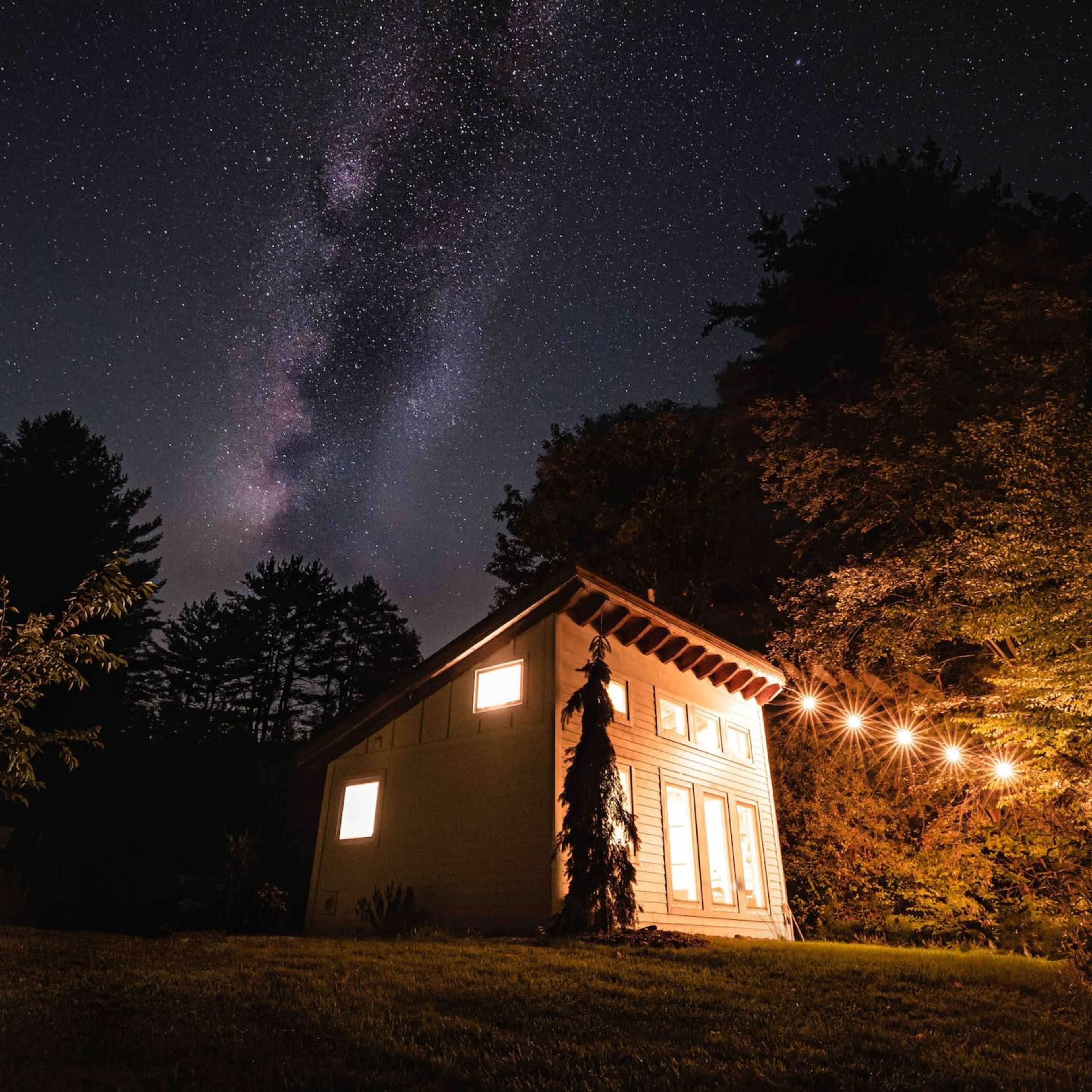A small house is illuminated by warm lights under a starry night sky with the Milky Way visible above.