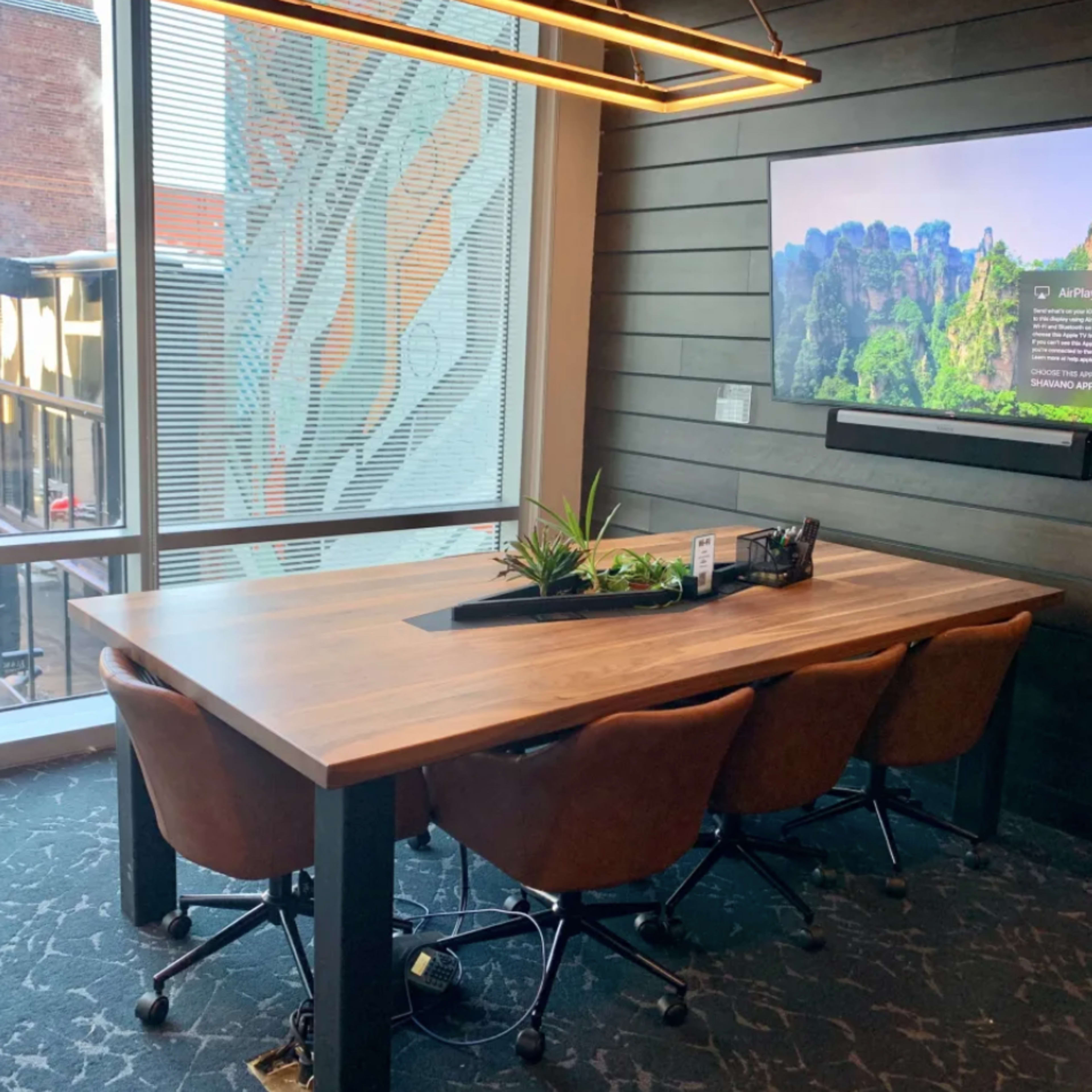 A conference table with four brown chairs is set against a window, featuring a television mounted on the wall displaying a nature scene.