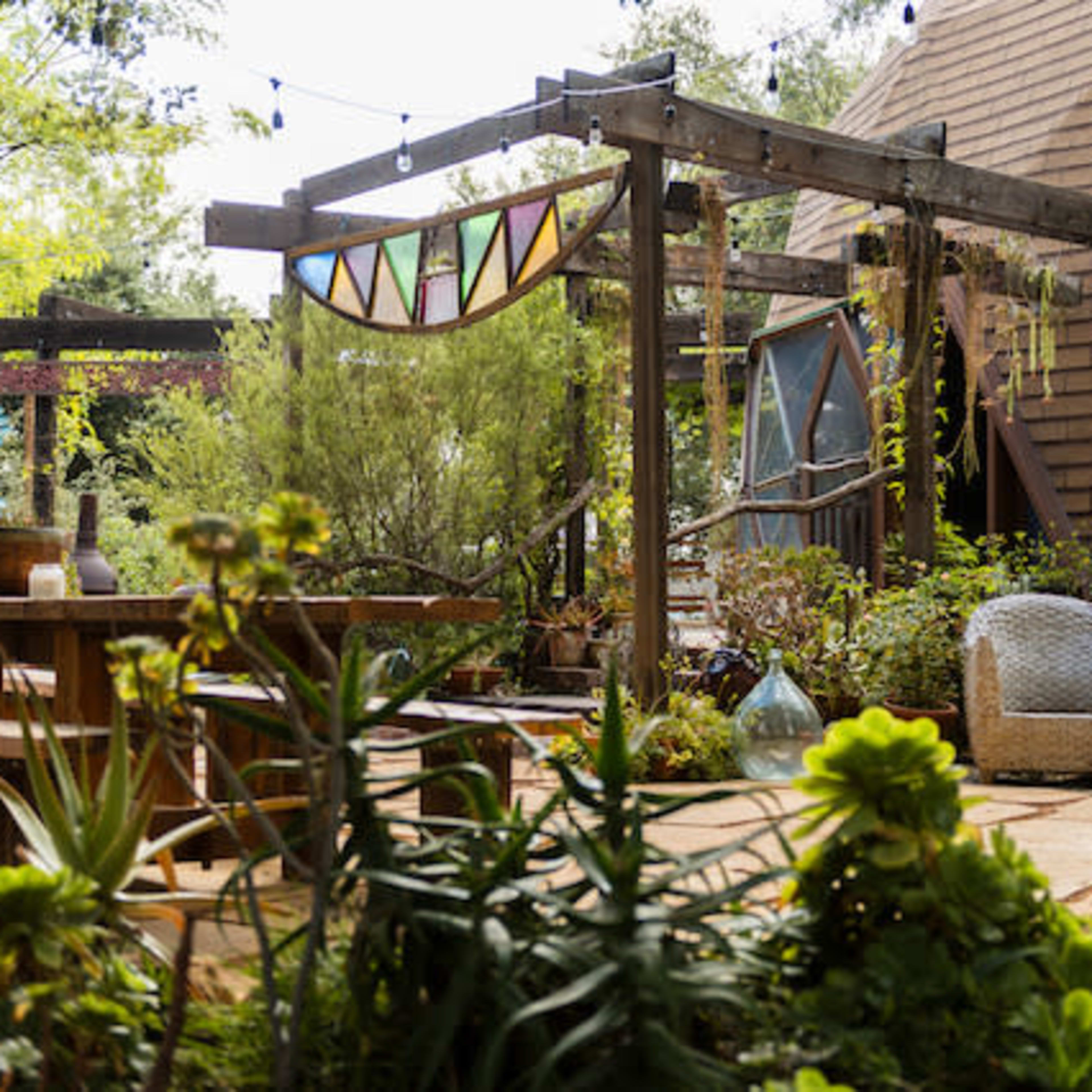 A landscaped garden area with wooden structures, a dining table, and decorative elements amid various plants and seating arrangements.