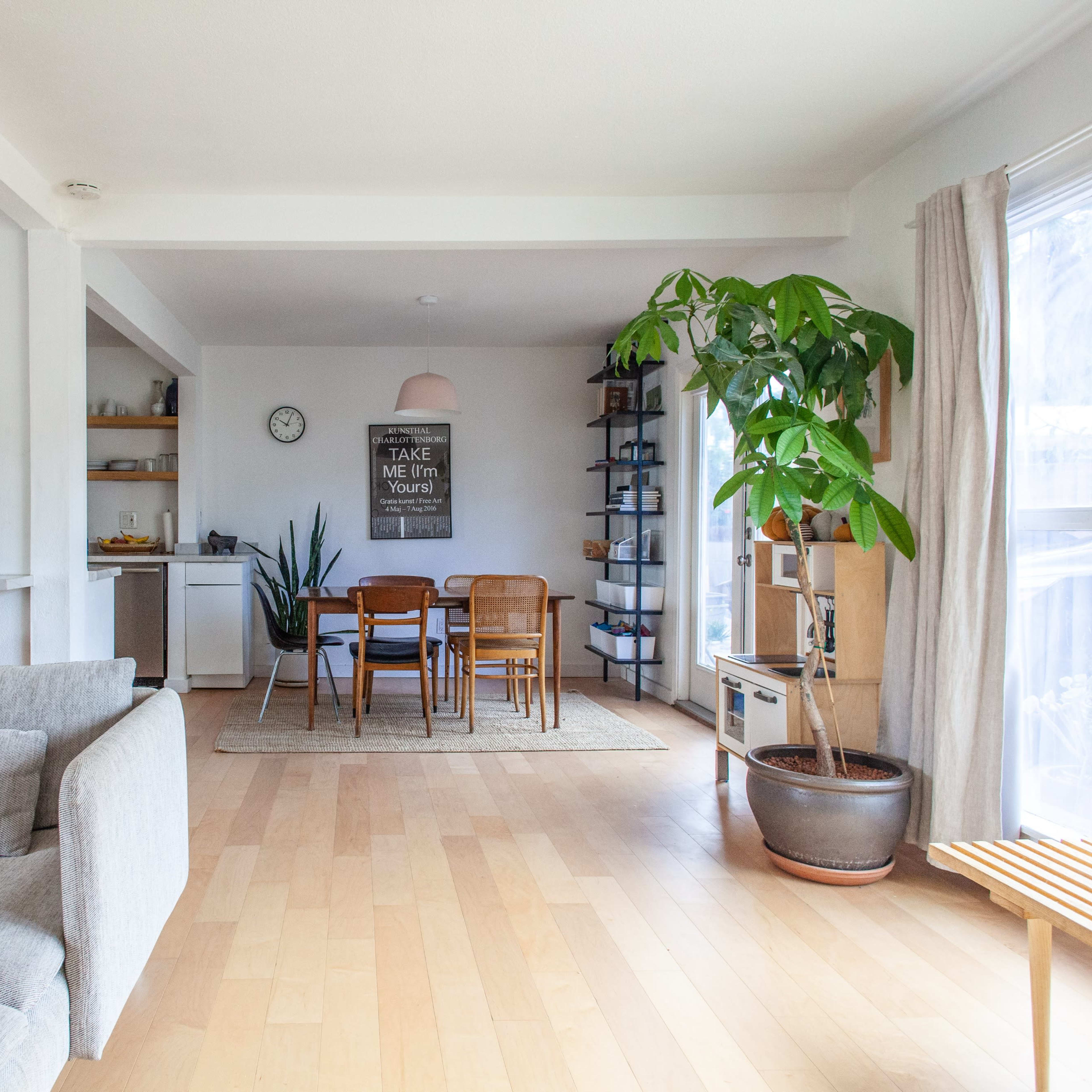 A modern, open-concept living space featuring a light-colored sofa, dining area with wooden furniture, and large windows allowing natural light.