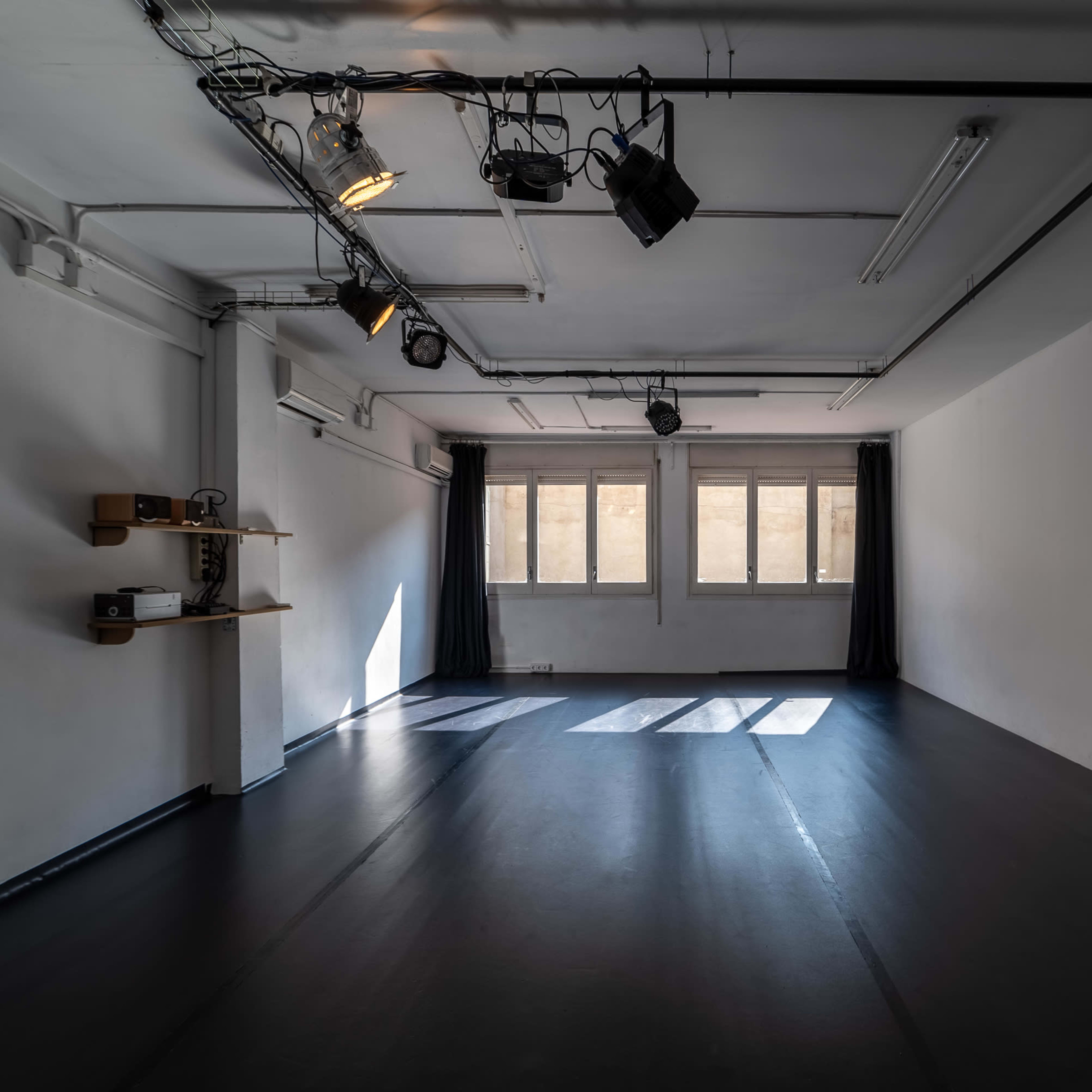 An empty, well-lit studio space with gray walls, black flooring, and daylight streaming through large windows.