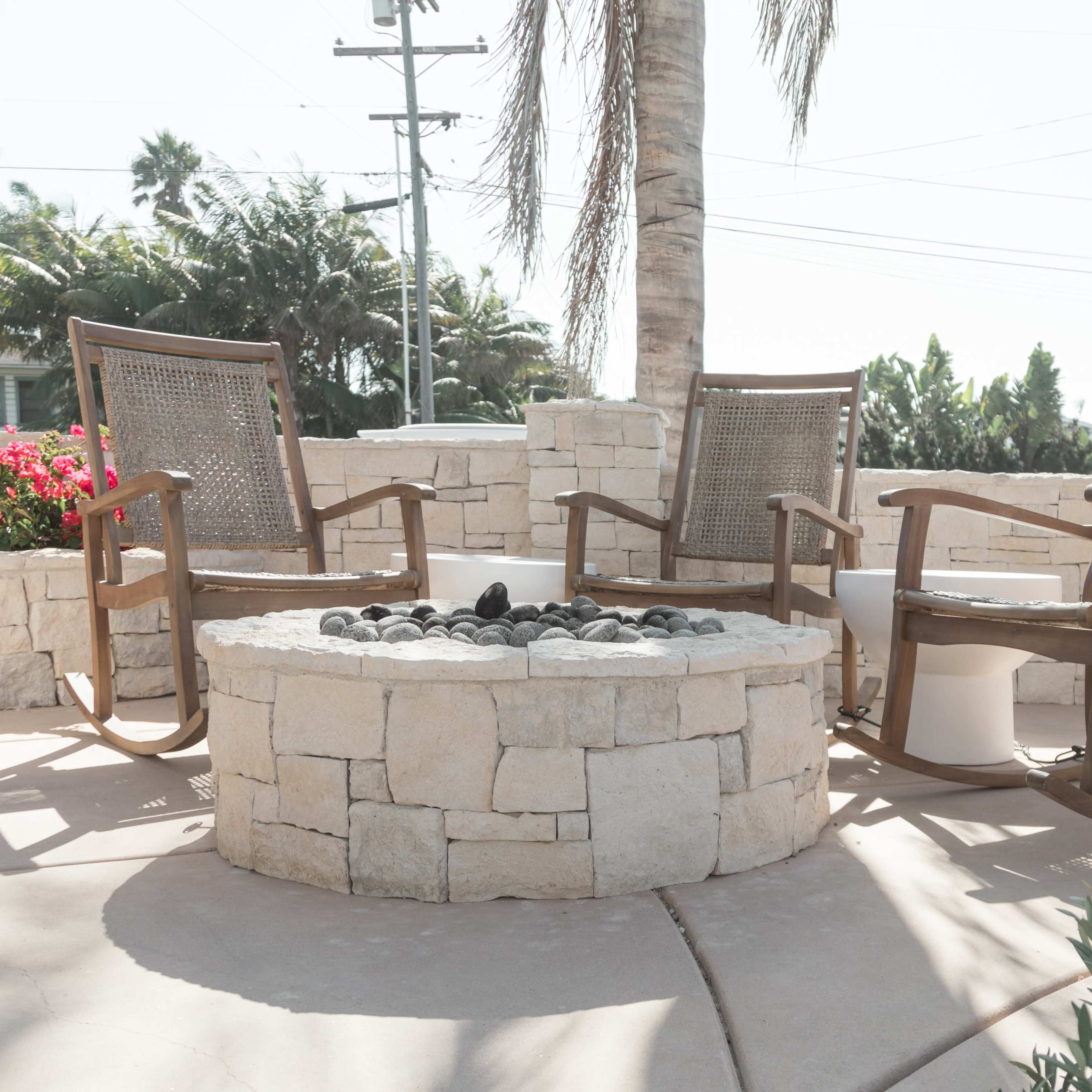 Three wooden chairs arranged around a circular stone fire pit, surrounded by plants and flowers in a sunny outdoor setting.