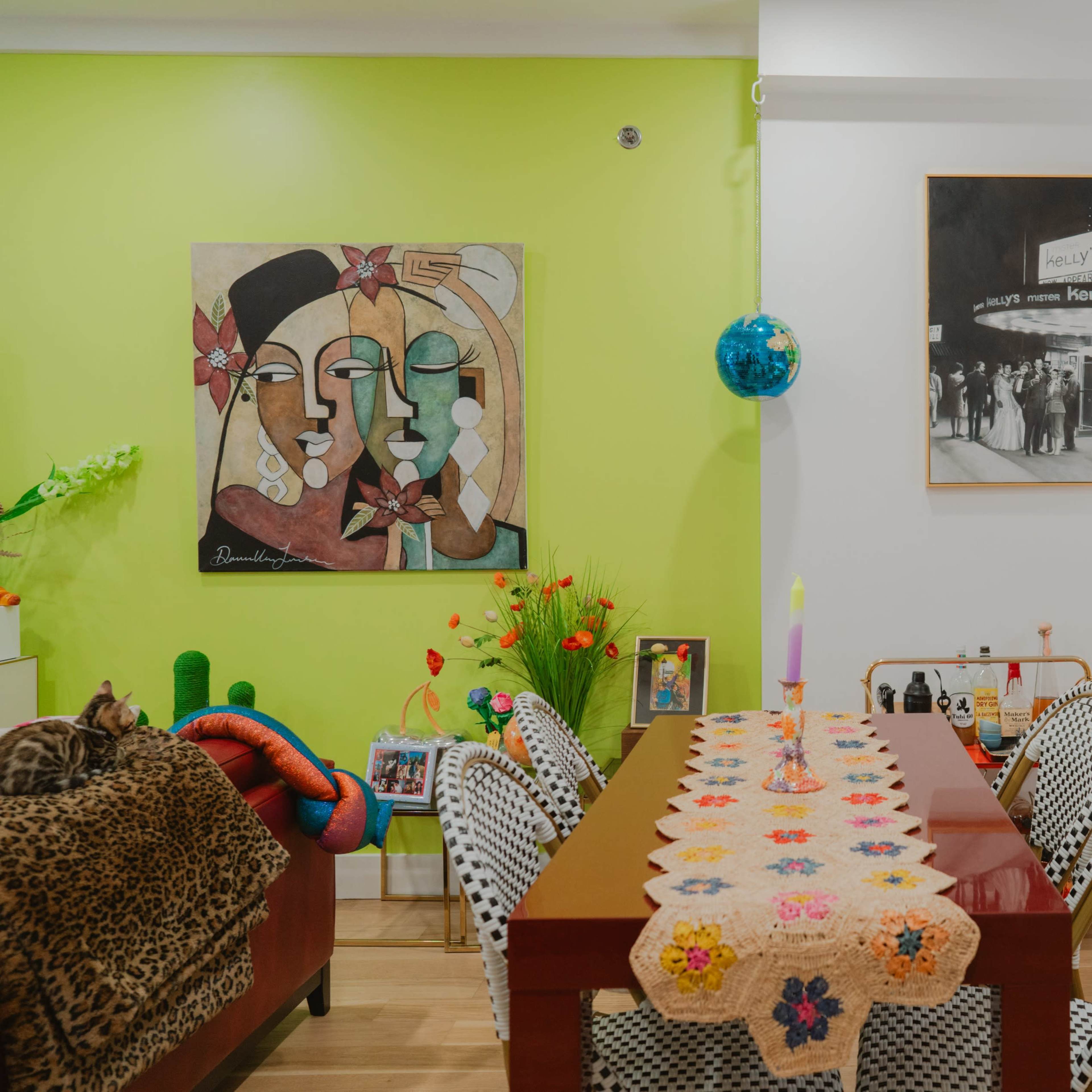A brightly colored room divided by a green wall, featuring a dining table with a colorful table runner, a mix of art pieces, and modern bar stools.