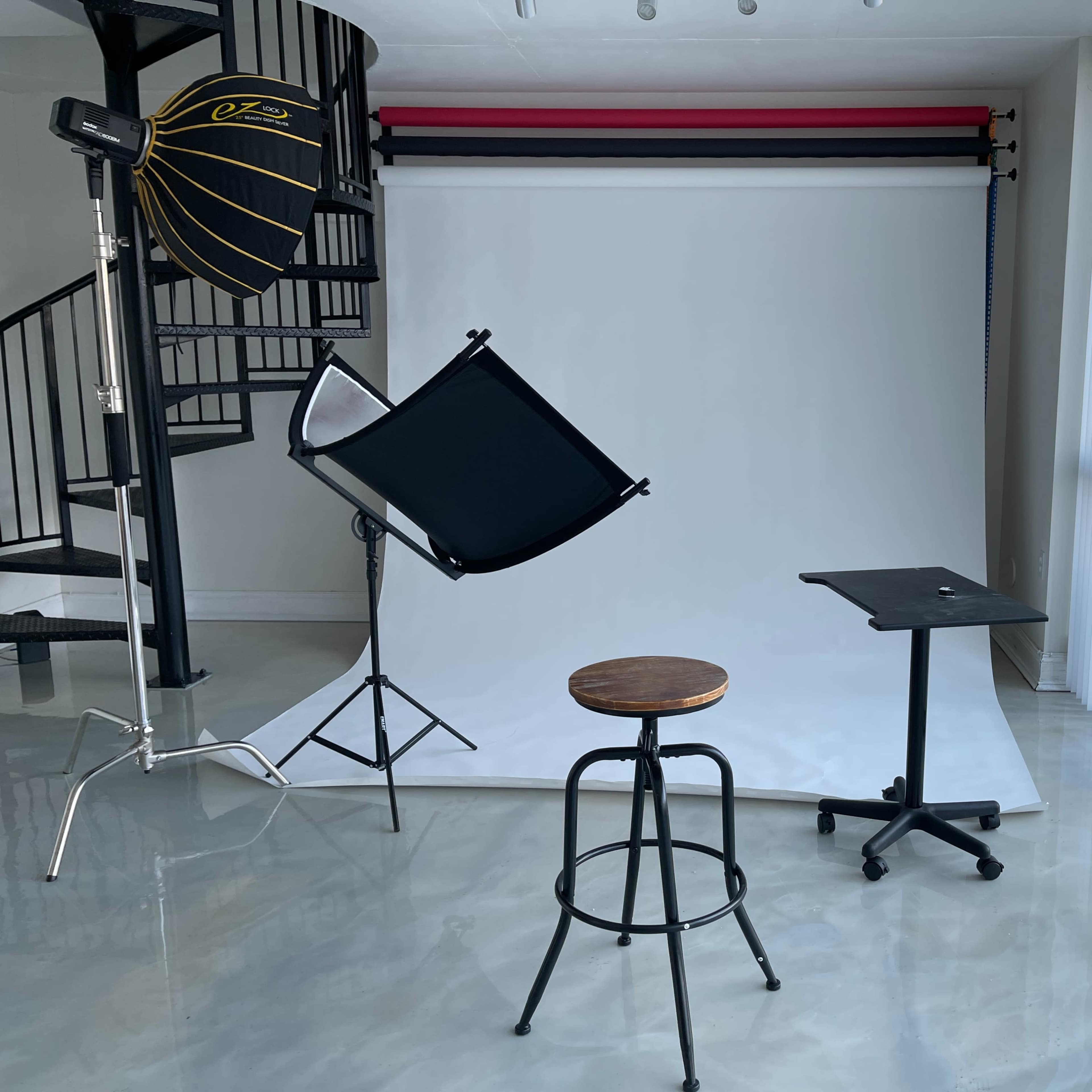 A photography studio features a white backdrop, a tall stool, a collapsible table, and lighting equipment.