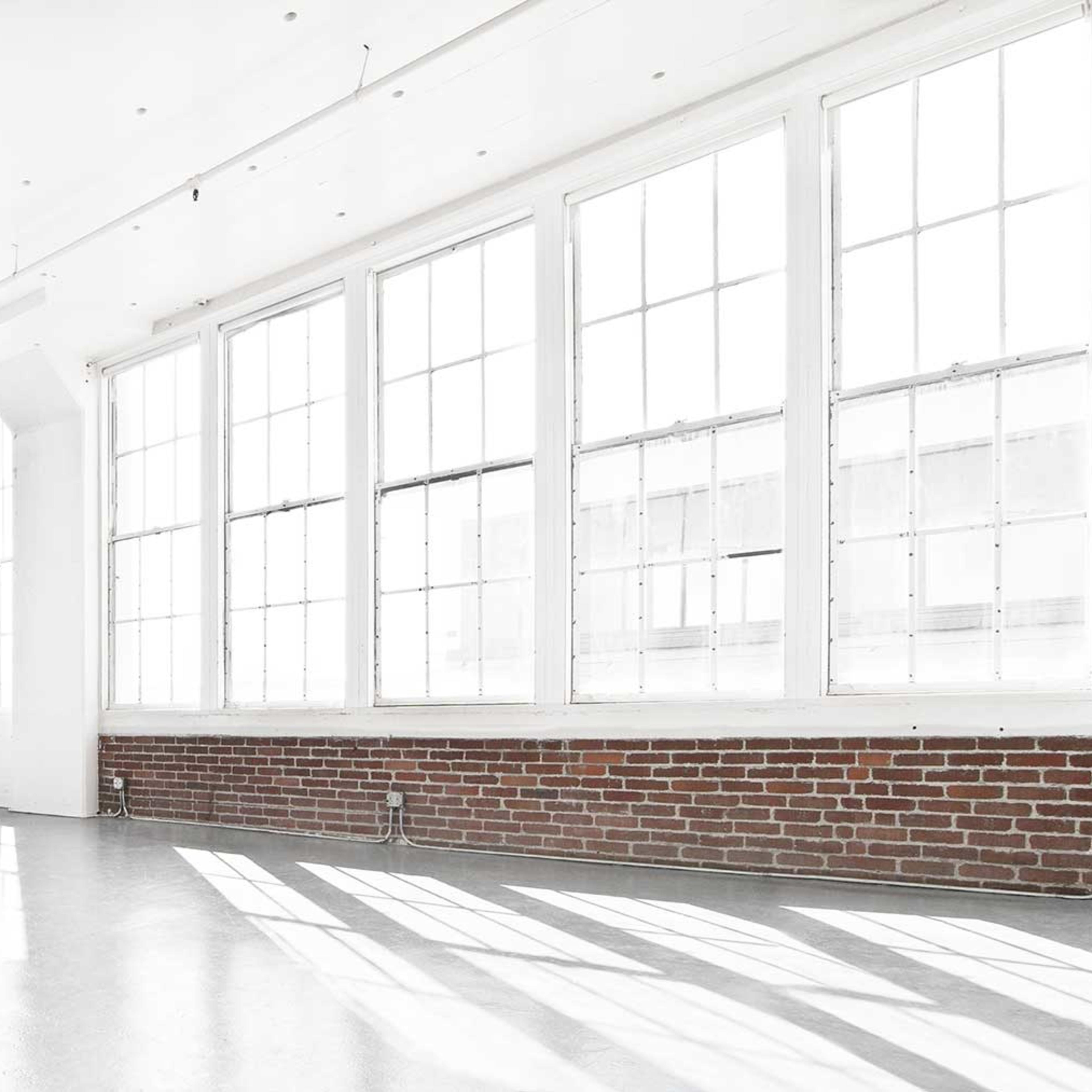 A spacious room with large windows allowing natural light to illuminate the white walls and floor, contrasted by a section of exposed brick.