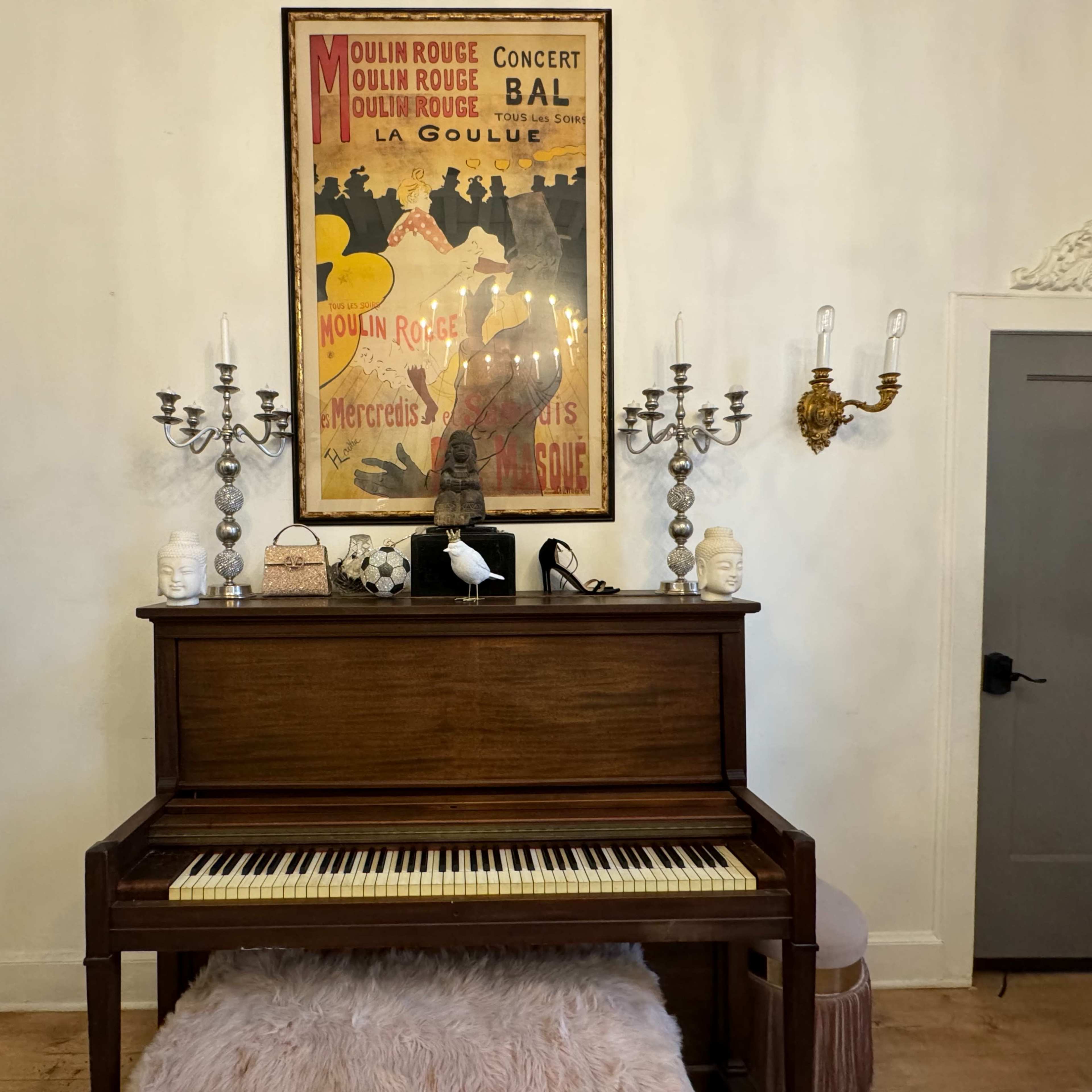 A wooden piano is positioned against a wall adorned with a vintage Moulin Rouge poster, flanked by decorative candelabras and a small statue.
