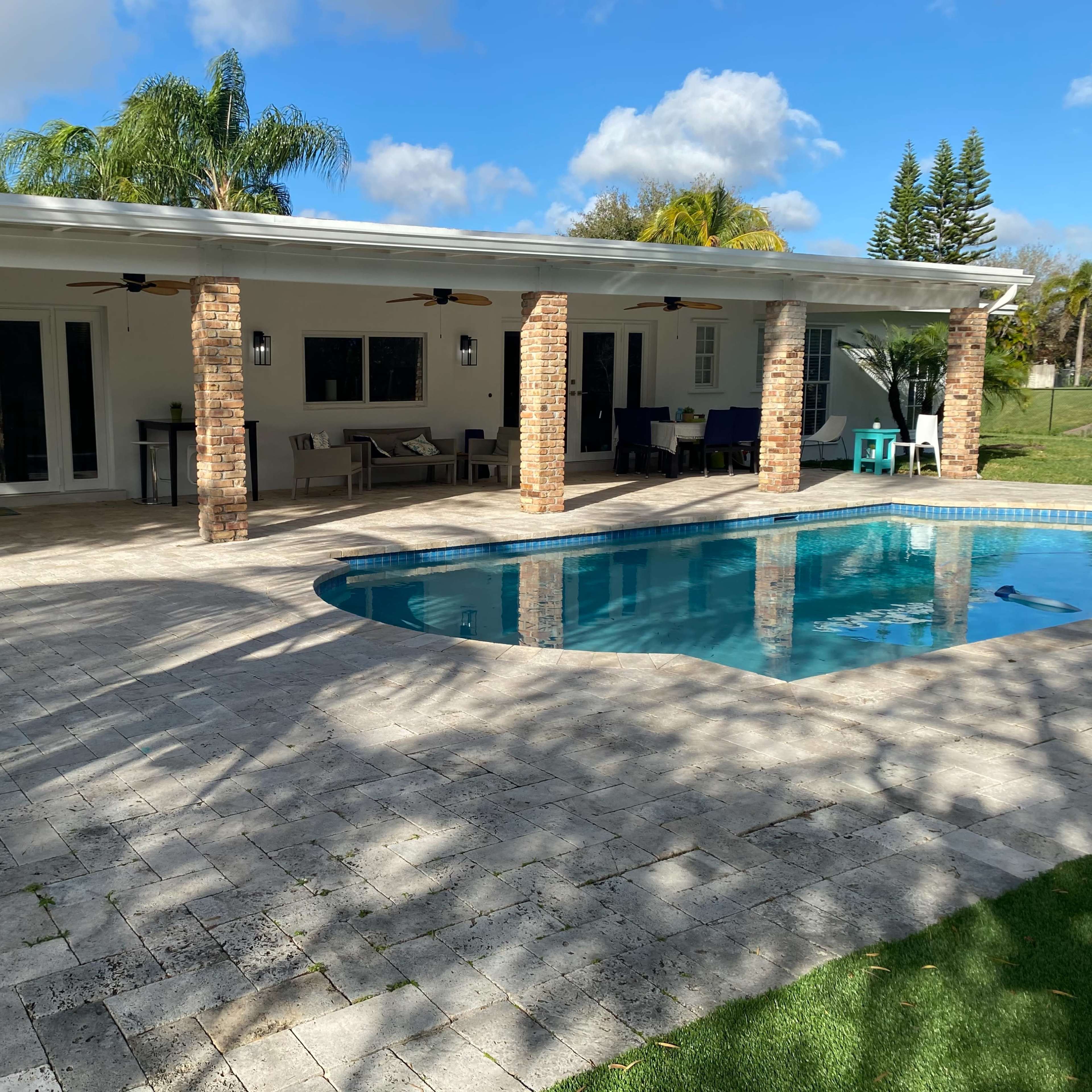 A modern outdoor space features a rectangular pool surrounded by a paved patio, with a covered sitting area and columns made of brick.