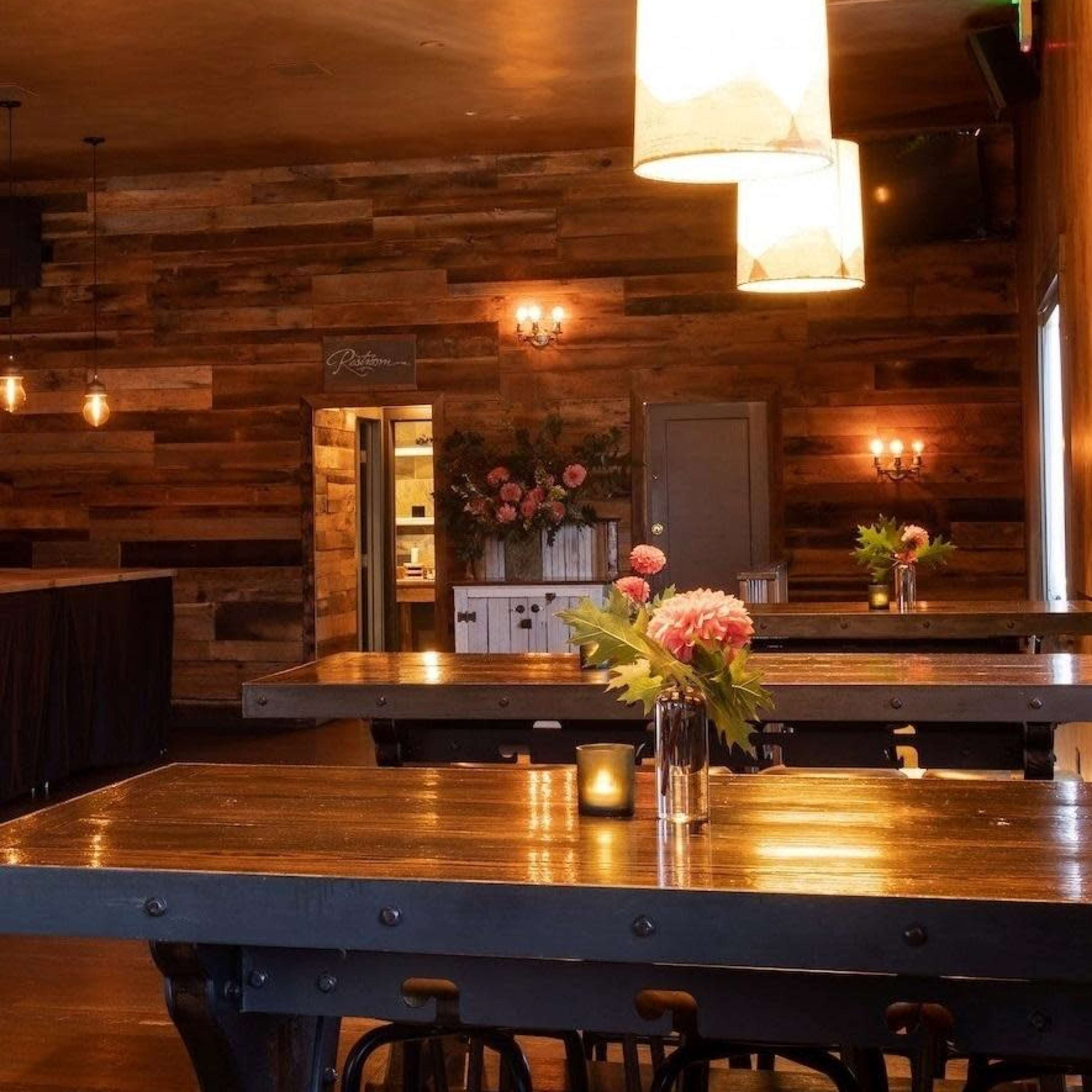 A cozy dining area features wooden tables, illuminated by pendant lights, with floral centerpieces on the tables and a wooden wall in the background.