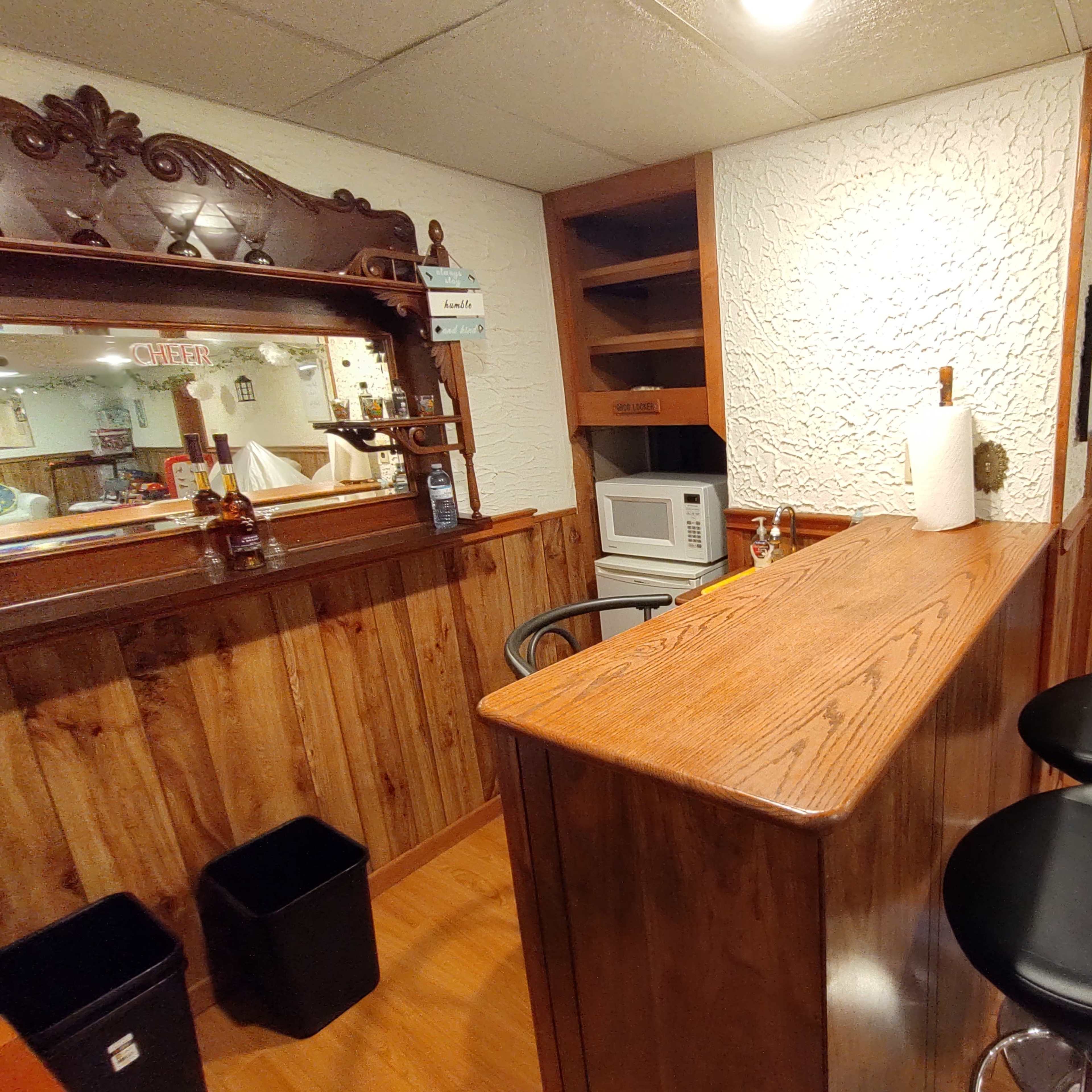 A small home bar area with a wooden counter, black bar stools, a microwave, and a mirror on the wall.