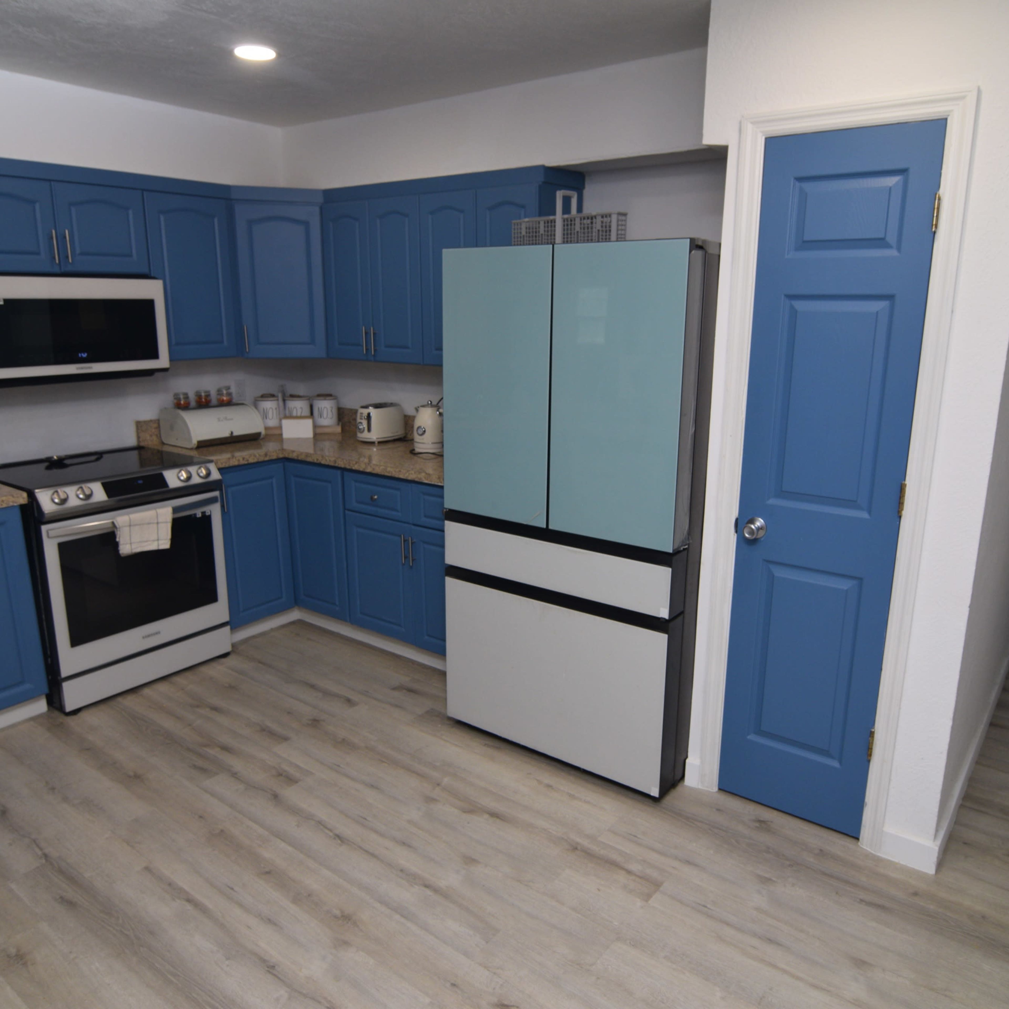 A kitchen with blue cabinets, a white stove, and a two-door refrigerator, featuring a light wood flooring and an adjoining hallway.