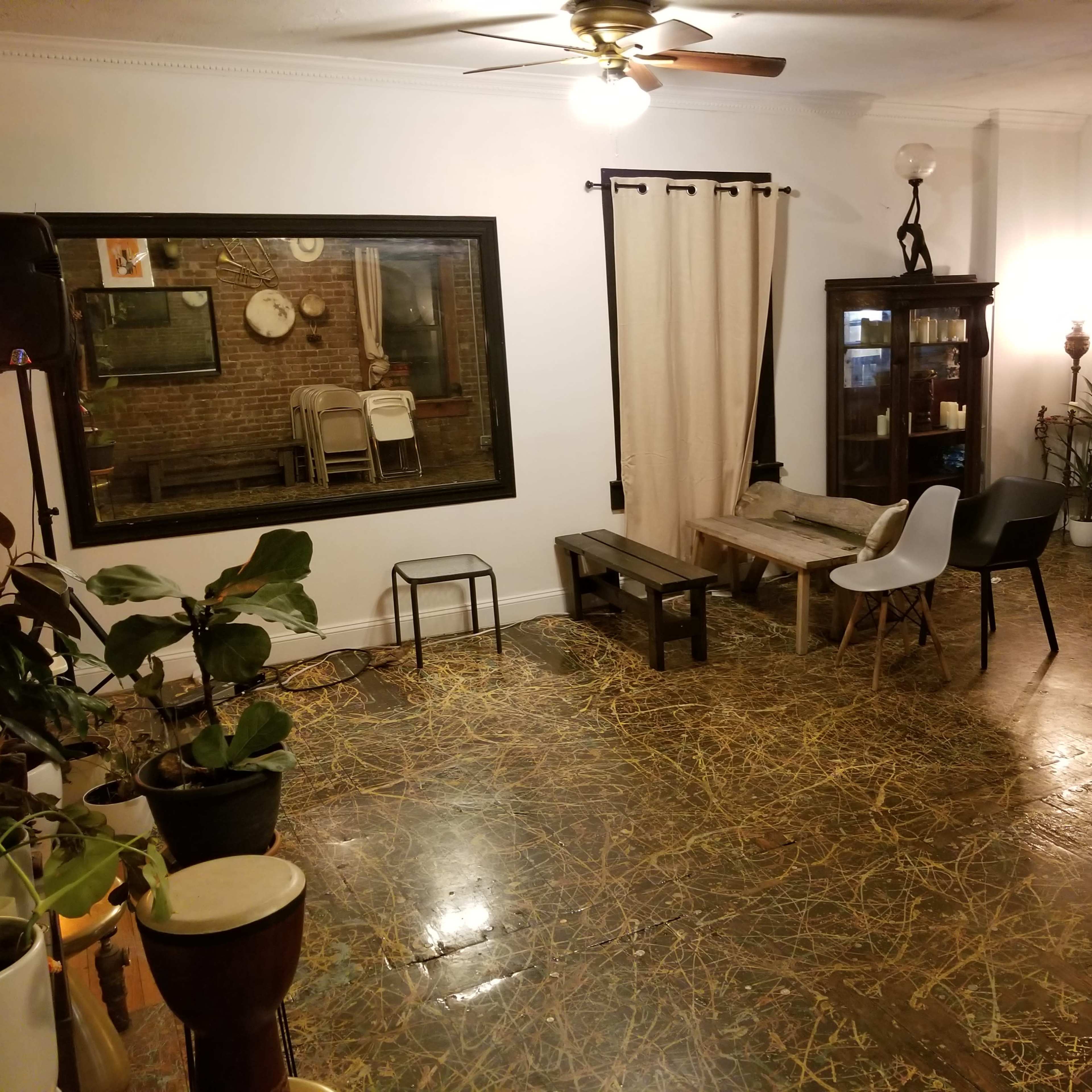 The interior of a room features wooden flooring, a large mirror, various chairs, a curtain, and a display cabinet against a brick wall.