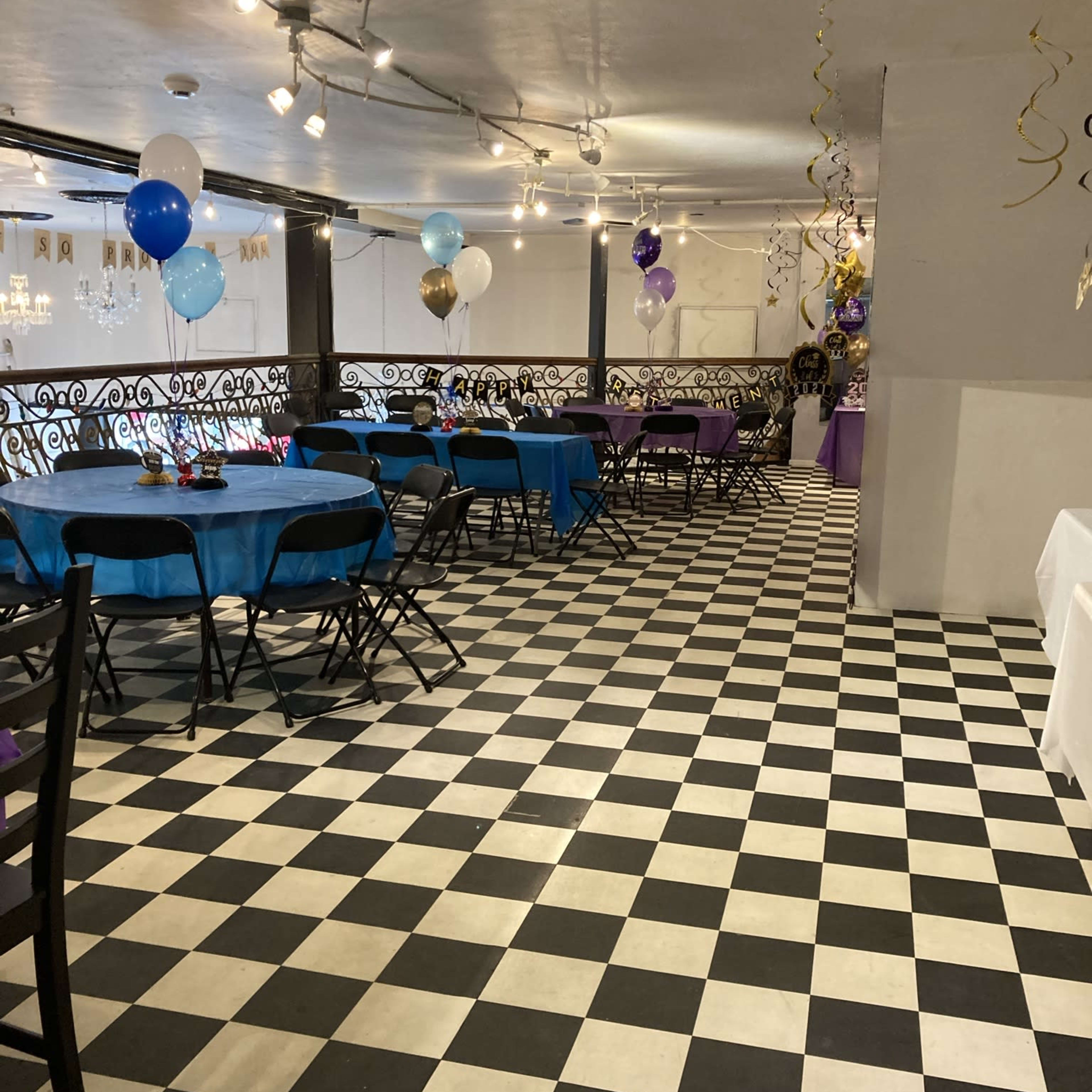A large indoor space set up for a celebration, featuring black and white checkered flooring, blue and purple table covers, and colorful balloons.