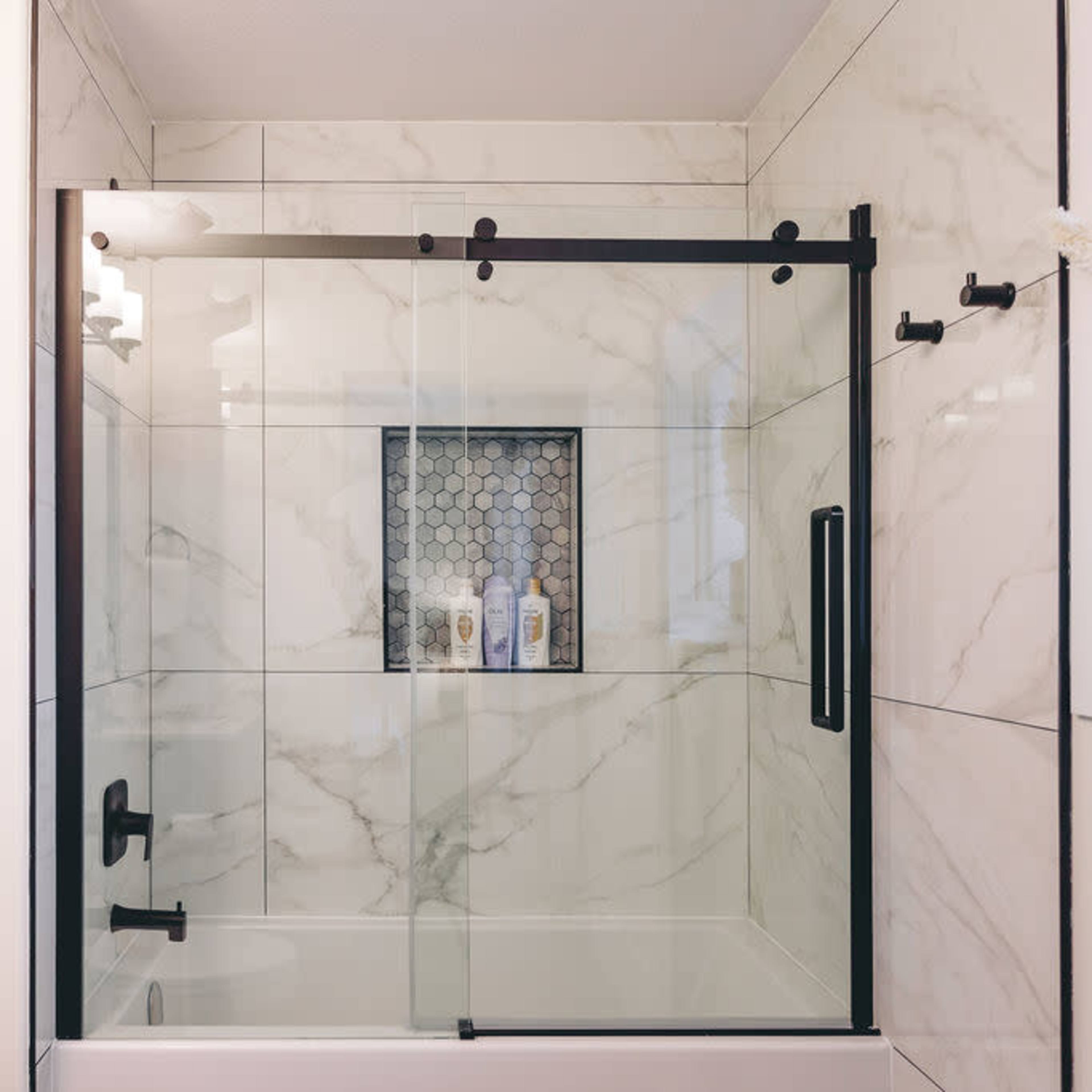 A modern glass shower enclosure with a black frame, set against white marble walls and a decorative tile niche holding several bottles.