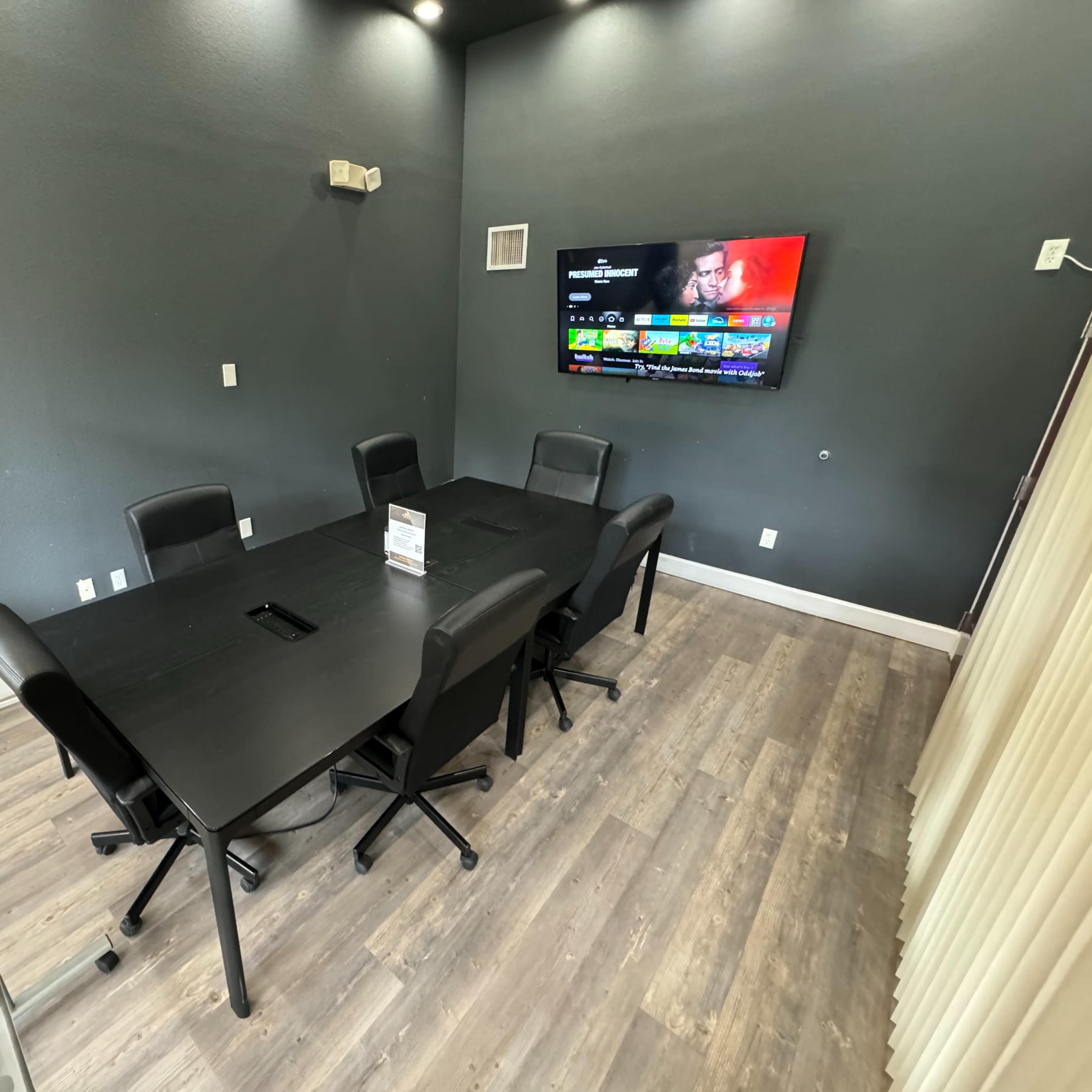A conference room features a black rectangular table surrounded by six chairs and a wall-mounted TV.