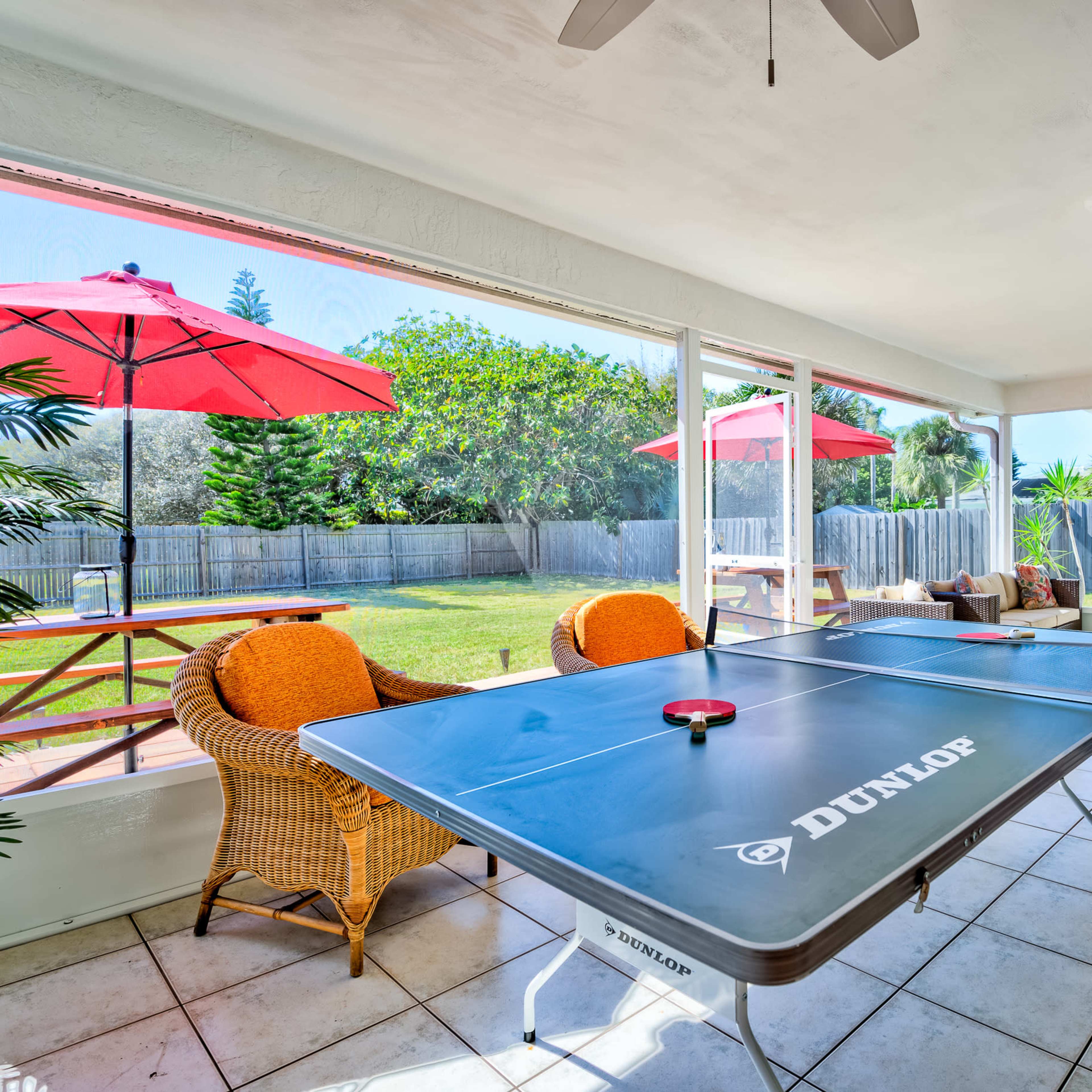 A ping pong table is set up in a bright, open room overlooking a backyard with picnic tables and red umbrellas.